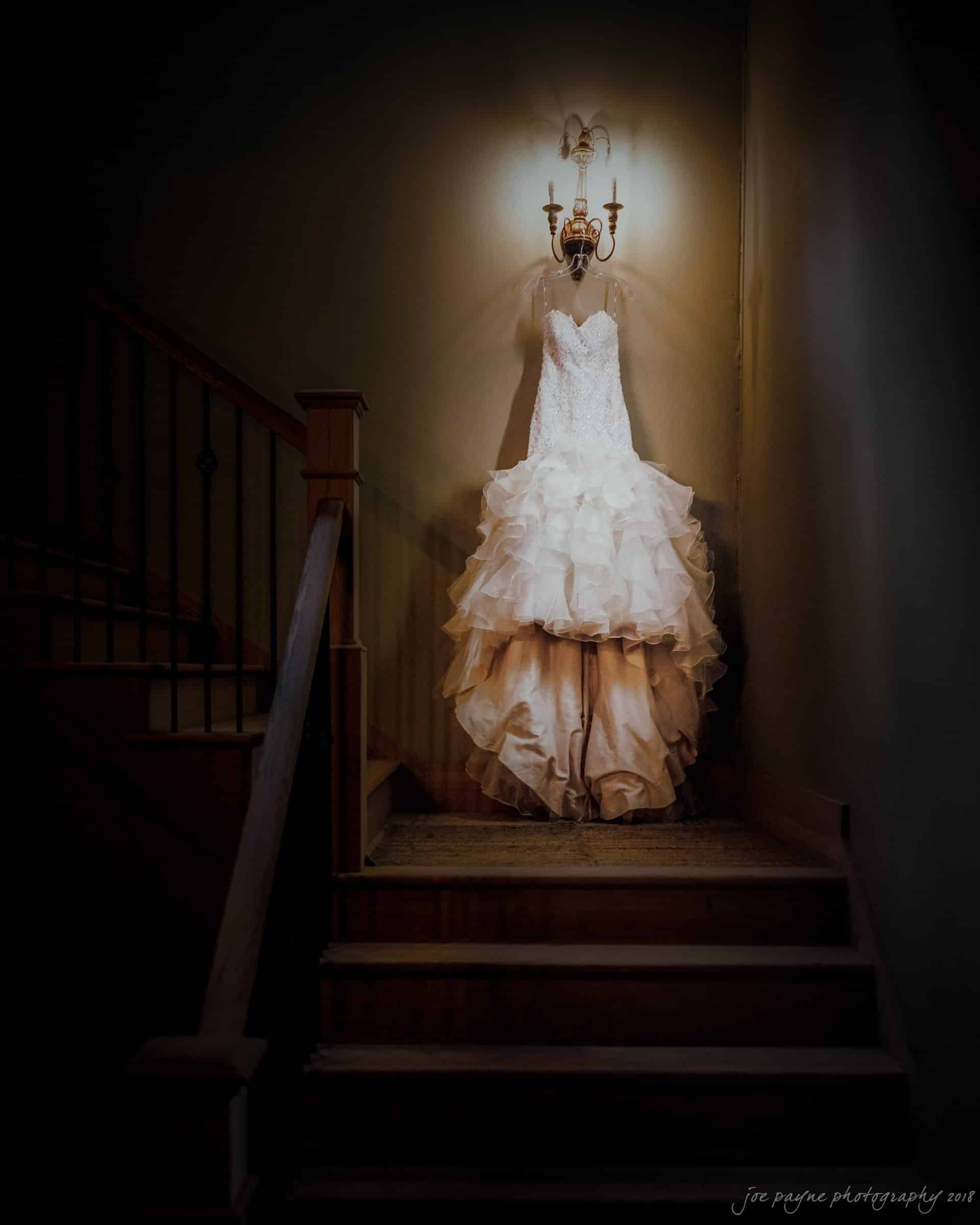 pavilion at carriage farm wedding - ashley & devon A ruffled ivory wedding gown is elegantly hung on a wall sconce in a dimly lit stairwell. The soft lighting casts warm shadows on the dress, highlighting its intricate design and creating a romantic atmosphere.