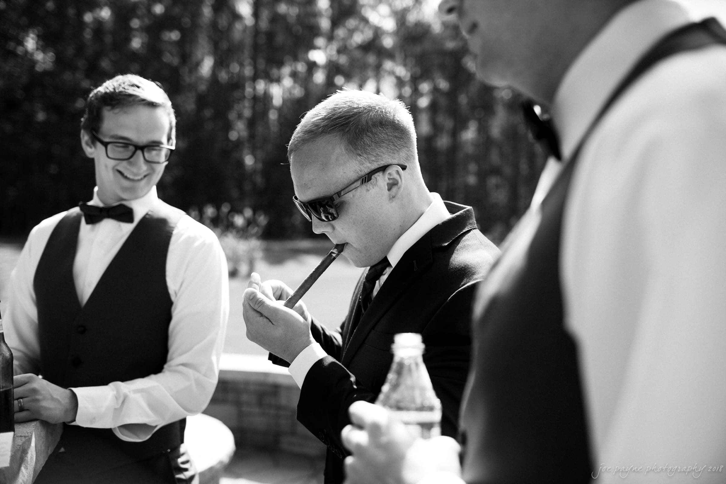 pavilion at carriage farm wedding - ashley & devon A black and white photo shows three men in formal attire. One man is lighting a cigar, wearing sunglasses. Another holds a water bottle, and the third, smiling, wears glasses. Trees are visible in the background.