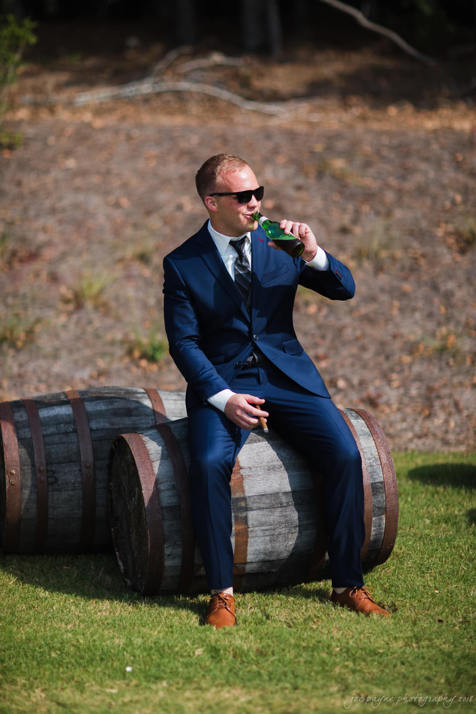 pavilion at carriage farm wedding - ashley & devon A man in a navy suit and sunglasses sits on wooden barrels outdoors, sipping from a green bottle. Sunny day with grass and a sloped dirt area in the background.