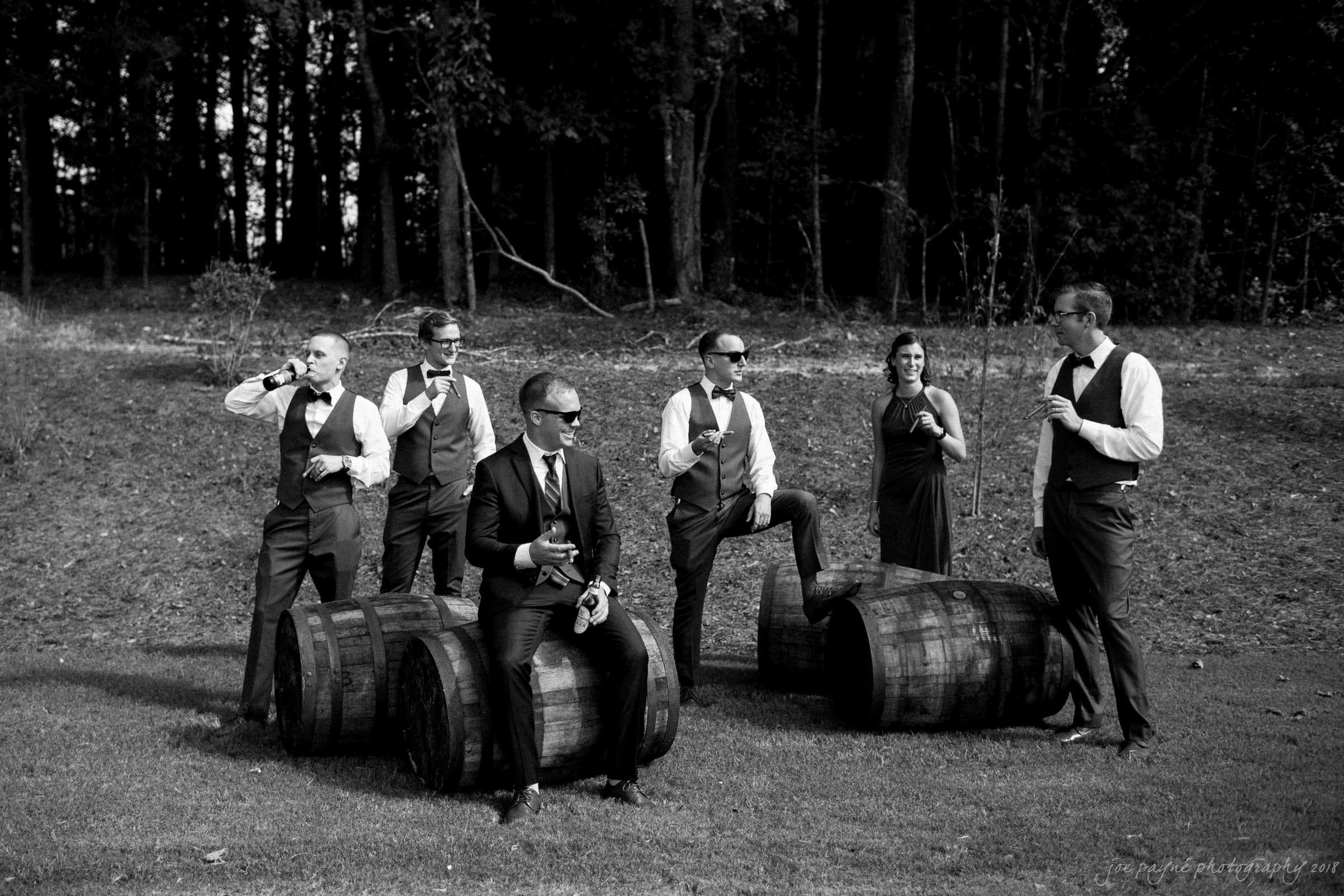 pavilion at carriage farm wedding - ashley & devon A black-and-white photo of a group of people in formal attire. They pose outdoors on grassy terrain with wooden barrels. Some are holding drinks. A forested area is visible in the background.