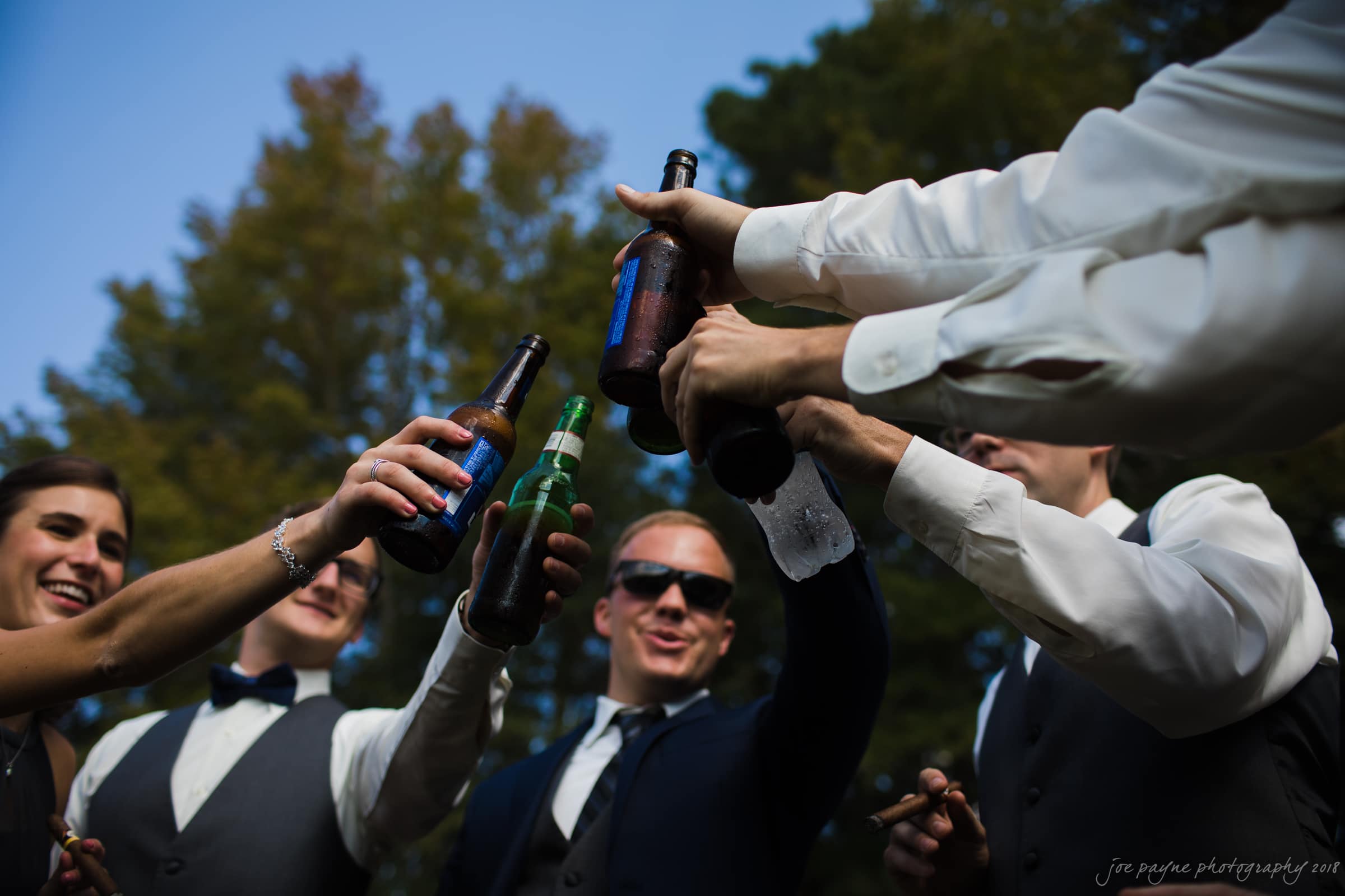 pavilion at carriage farm wedding - ashley & devon A group of individuals dressed in formal attire, raising beer bottles in a joyful outdoor toast. The focus is on their hands and bottles against a backdrop of trees and blue sky.
