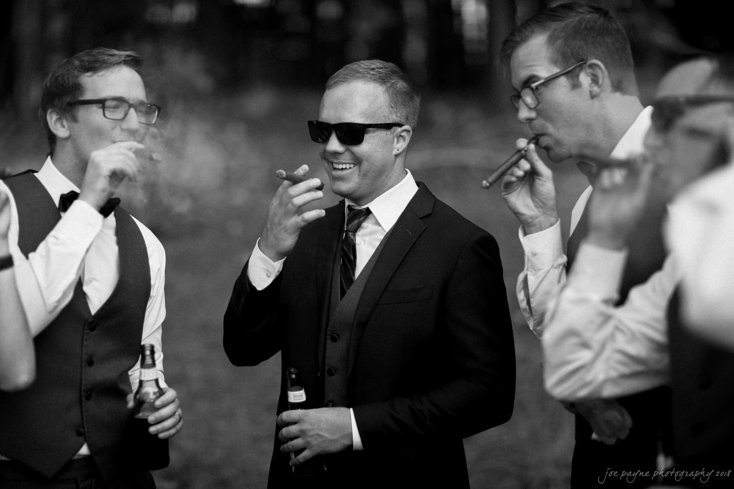 pavilion at carriage farm wedding - ashley & devon Four men in suits and sunglasses enjoy cigars and drinks outdoors. One man laughs while the others chat, creating a relaxed, celebratory atmosphere.