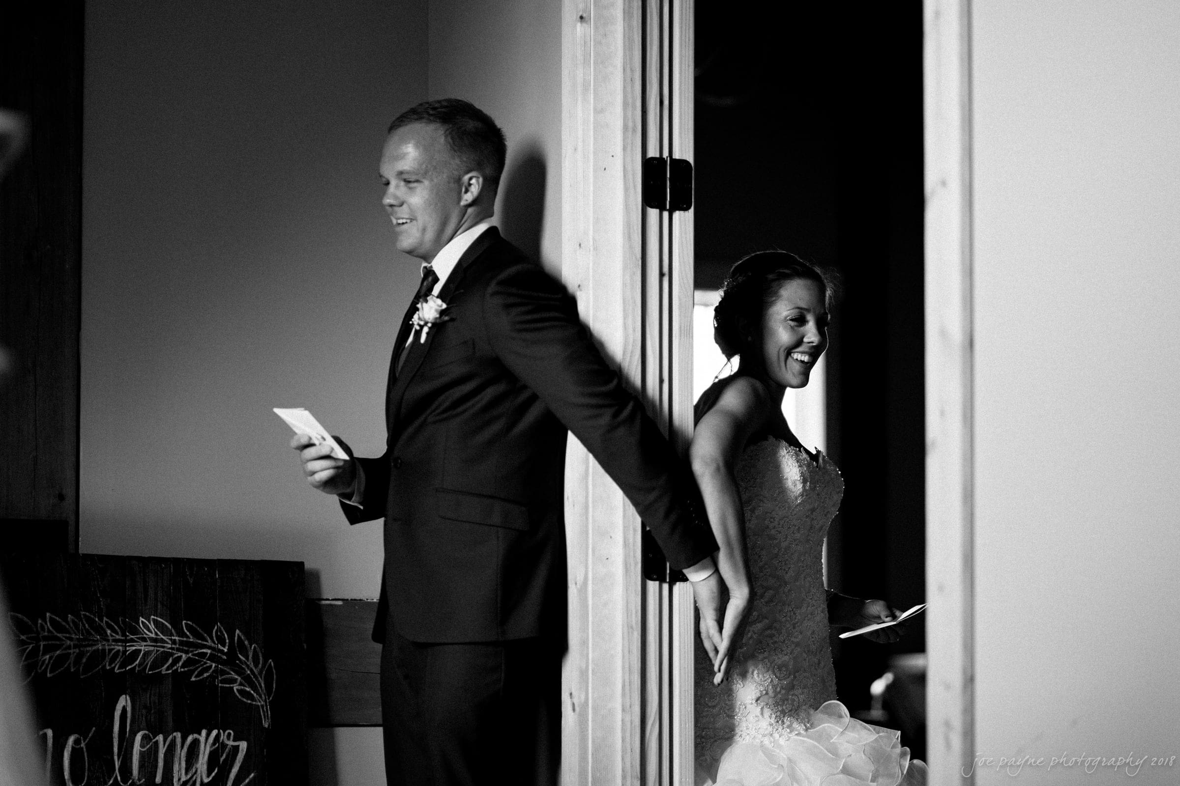 pavilion at carriage farm wedding - ashley & devon A bride and groom stand on opposite sides of a door, holding hands. Both are smiling, with the groom holding a note. The bride looks joyful in her wedding dress, while the groom is in a suit. The scene is captured in black and white.