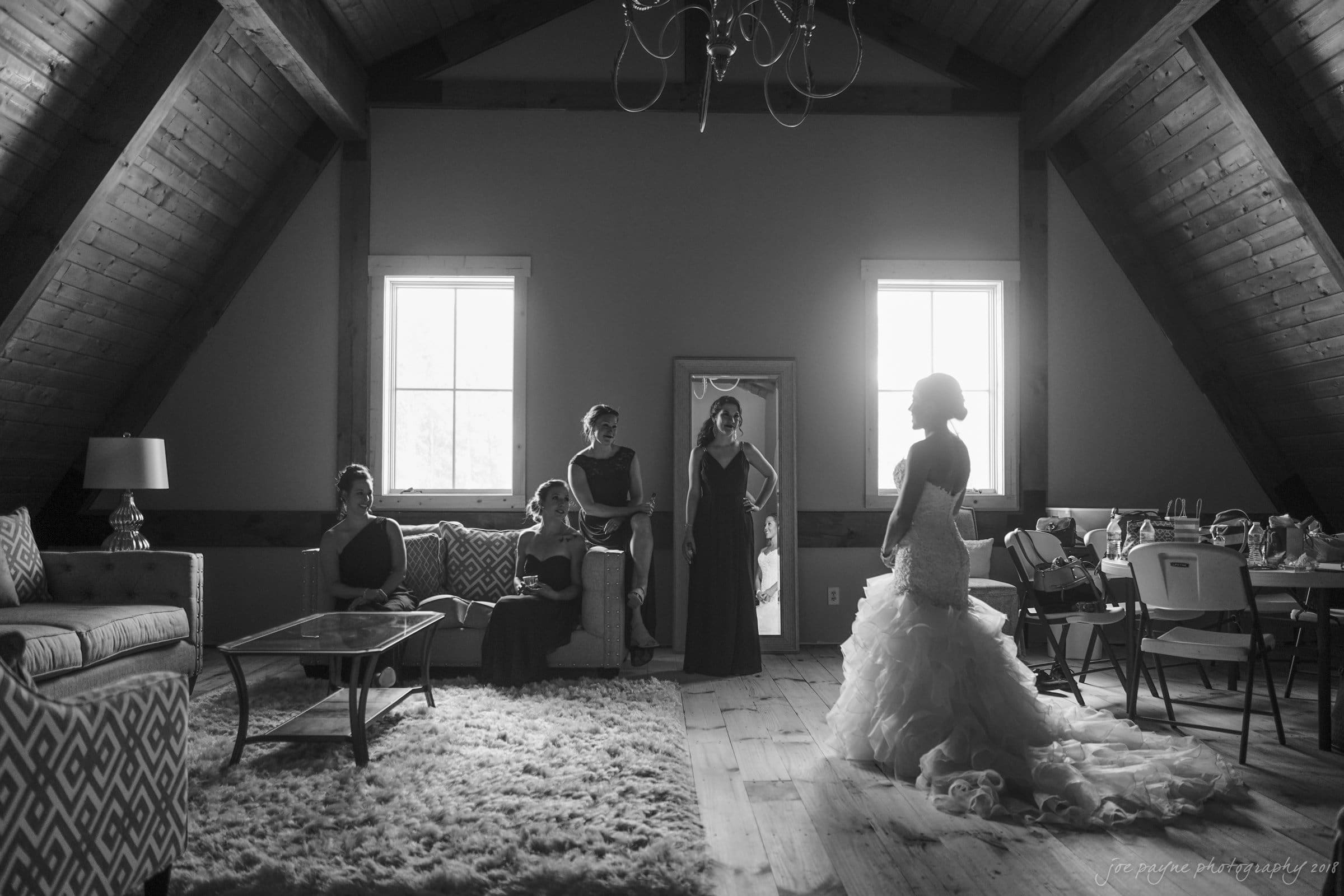 pavilion at carriage farm wedding - ashley & devon A black and white photo of a bride in her wedding dress standing in a room with bridesmaids. The room has wooden beams, a chandelier, and modern furniture. Natural light streams through two windows, casting soft shadows.