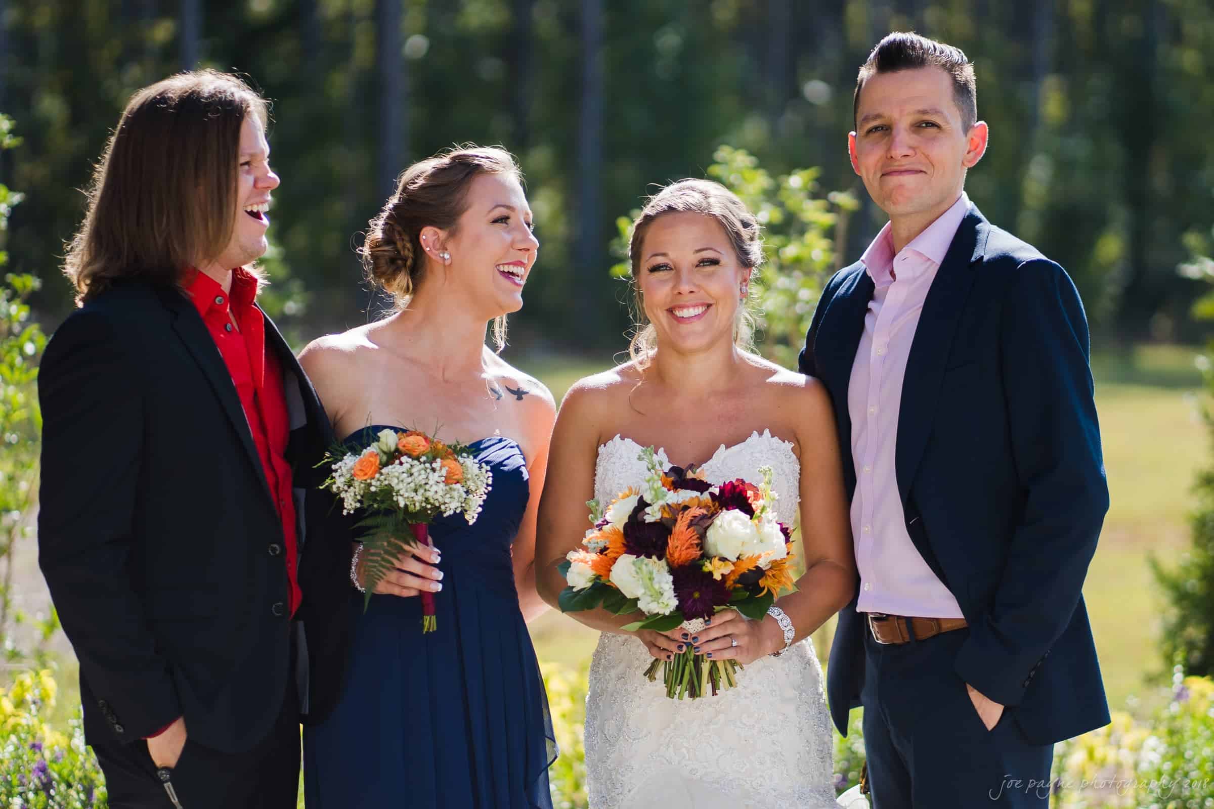 pavilion at carriage farm wedding - ashley & devon A bride in a white gown stands smiling with a bouquet of flowers. Beside her, a woman in a blue dress holds another bouquet, and two men in suits smile in the sunlight, with trees in the background.
