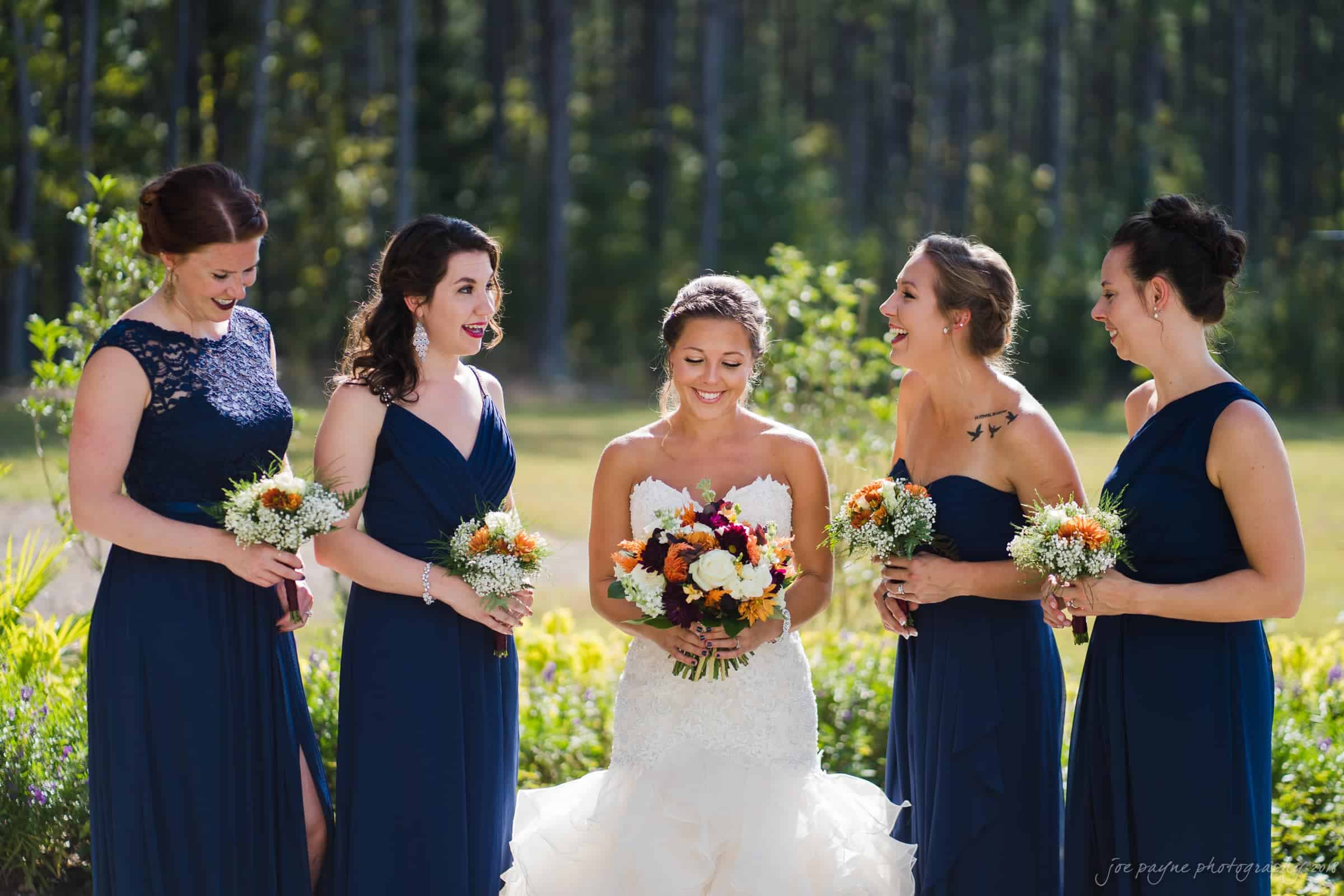 pavilion at carriage farm wedding - ashley & devon A bride in a white gown holding a bouquet stands with four bridesmaids in navy blue dresses, each holding small flower bouquets. They are outdoors with a forest backdrop, smiling and laughing together.
