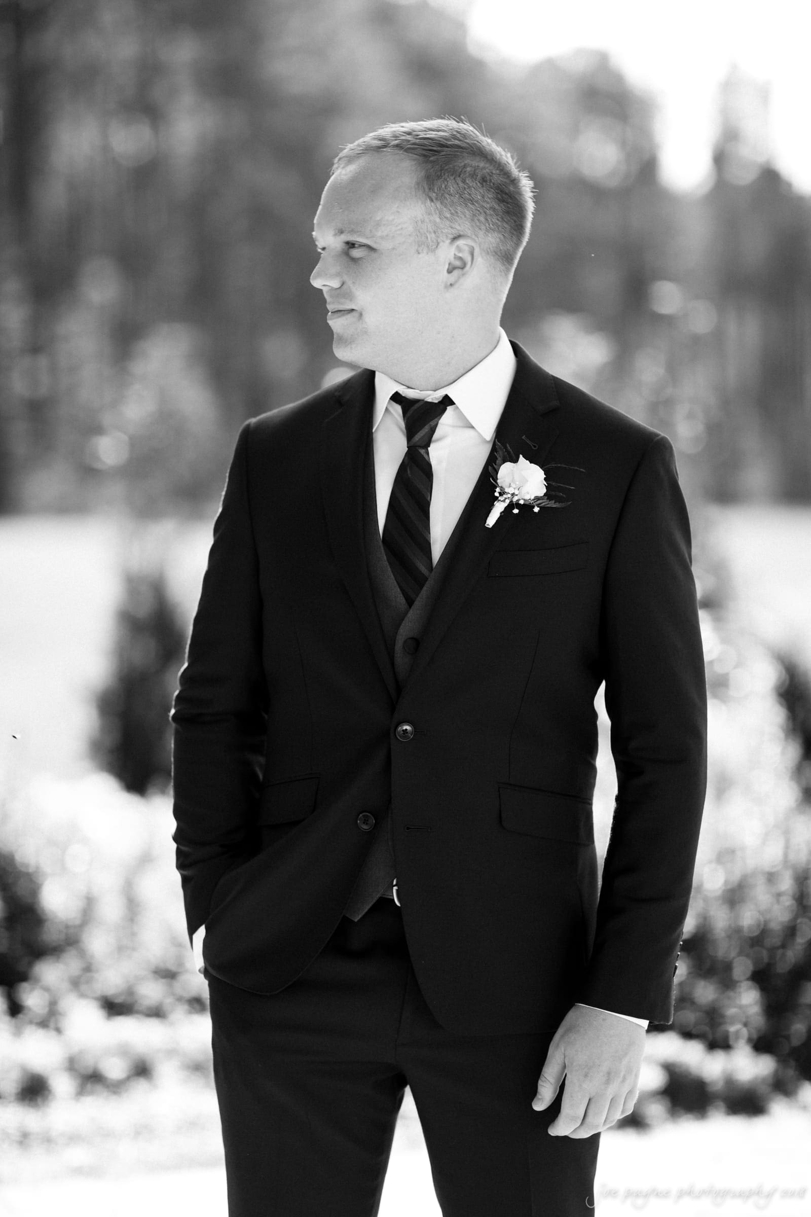 pavilion at carriage farm wedding - ashley & devon Black and white photo of a man in a suit standing outdoors. He is looking to the side, with one hand in his pocket. The background is blurred, featuring trees and greenery.