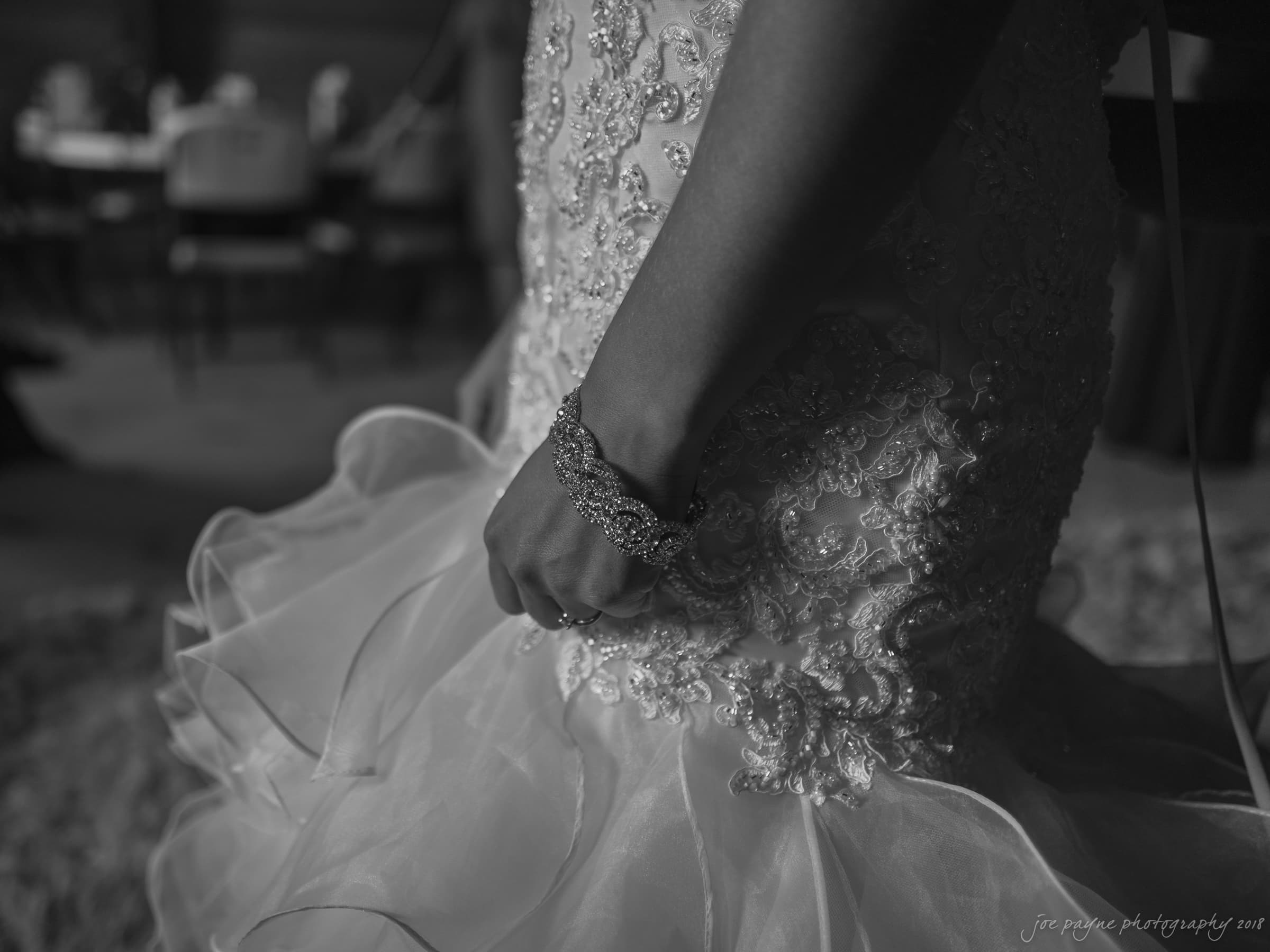pavilion at carriage farm wedding - ashley & devon A close-up black and white photo of a brides hand resting on her wedding dress. The dress features intricate lace and bead details. She wears a sparkling bracelet. The background is subtly blurred with chairs and tables visible.