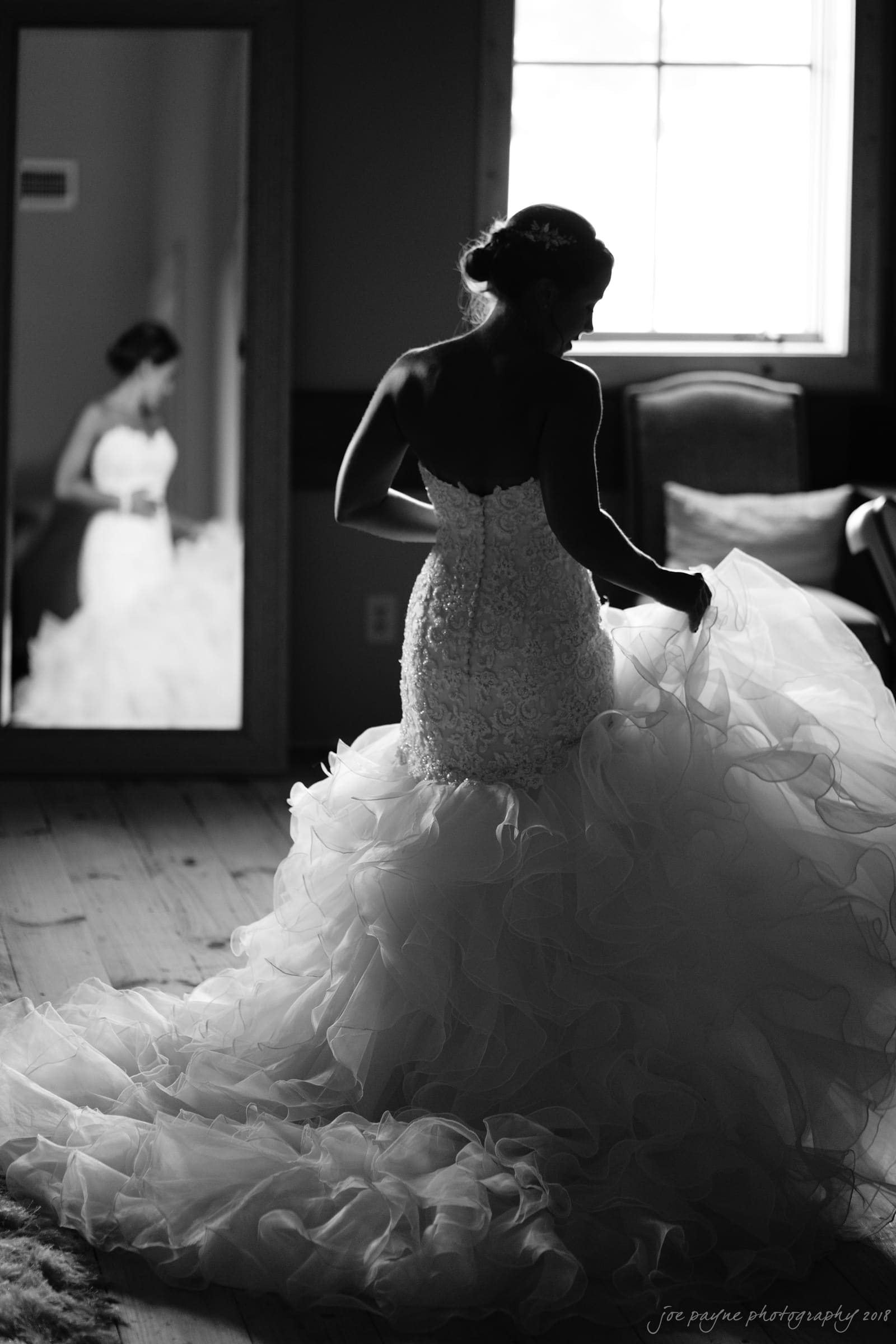 pavilion at carriage farm wedding - ashley & devon A bride in a flowing wedding dress stands before a large mirror in a softly lit room. The airy fabric cascades around her as she holds part of the skirt, with her reflection visible in the background.