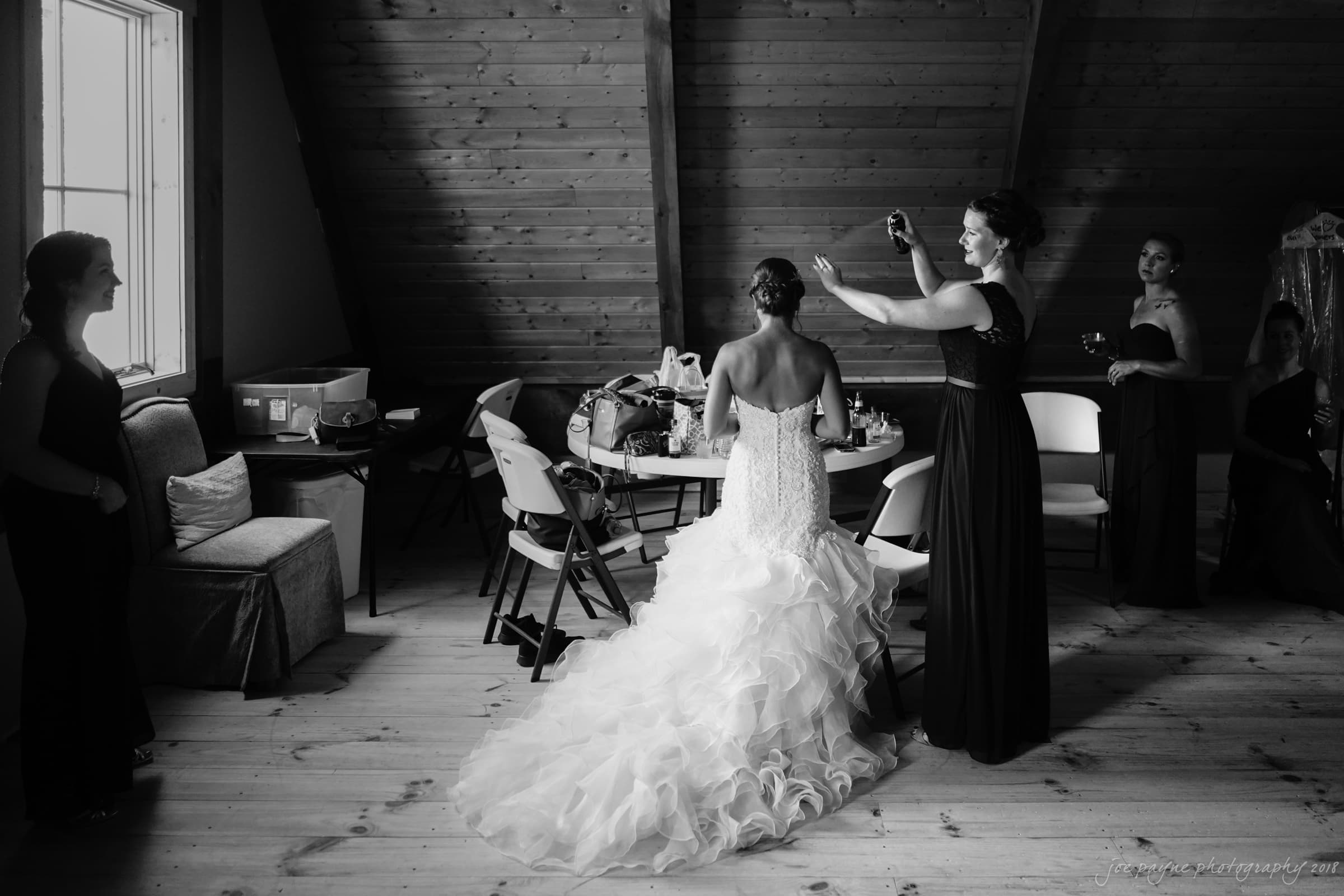 pavilion at carriage farm wedding - ashley & devon A black and white photo of a bride in a ruffled dress sitting inside a rustic wooden room. A bridesmaid assists with her hair. Other bridesmaids in dark gowns stand or sit nearby, chatting and preparing for the wedding.