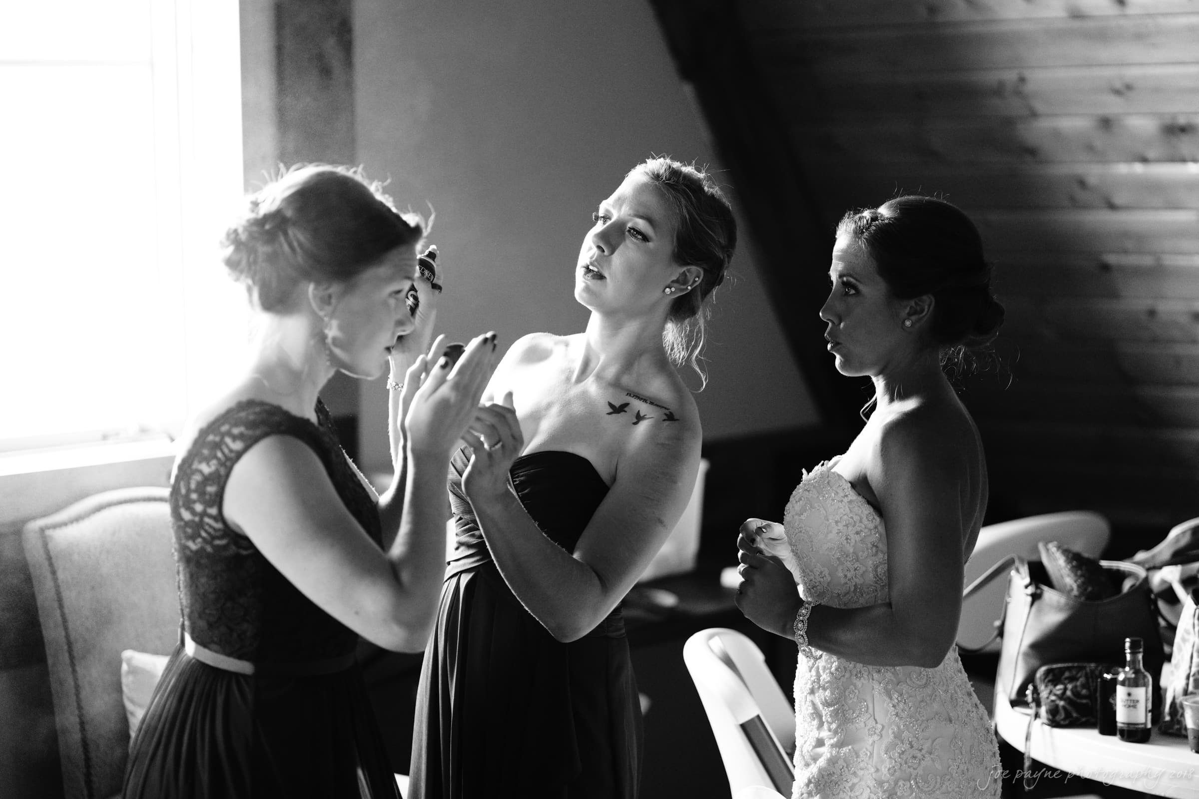 pavilion at carriage farm wedding - ashley & devon Three women in elegant dresses prepare in a softly lit room. One applies makeup, another adjusts her hair, and the third, in a wedding gown, observes. The setting has a cozy, rustic vibe with warm wooden walls.