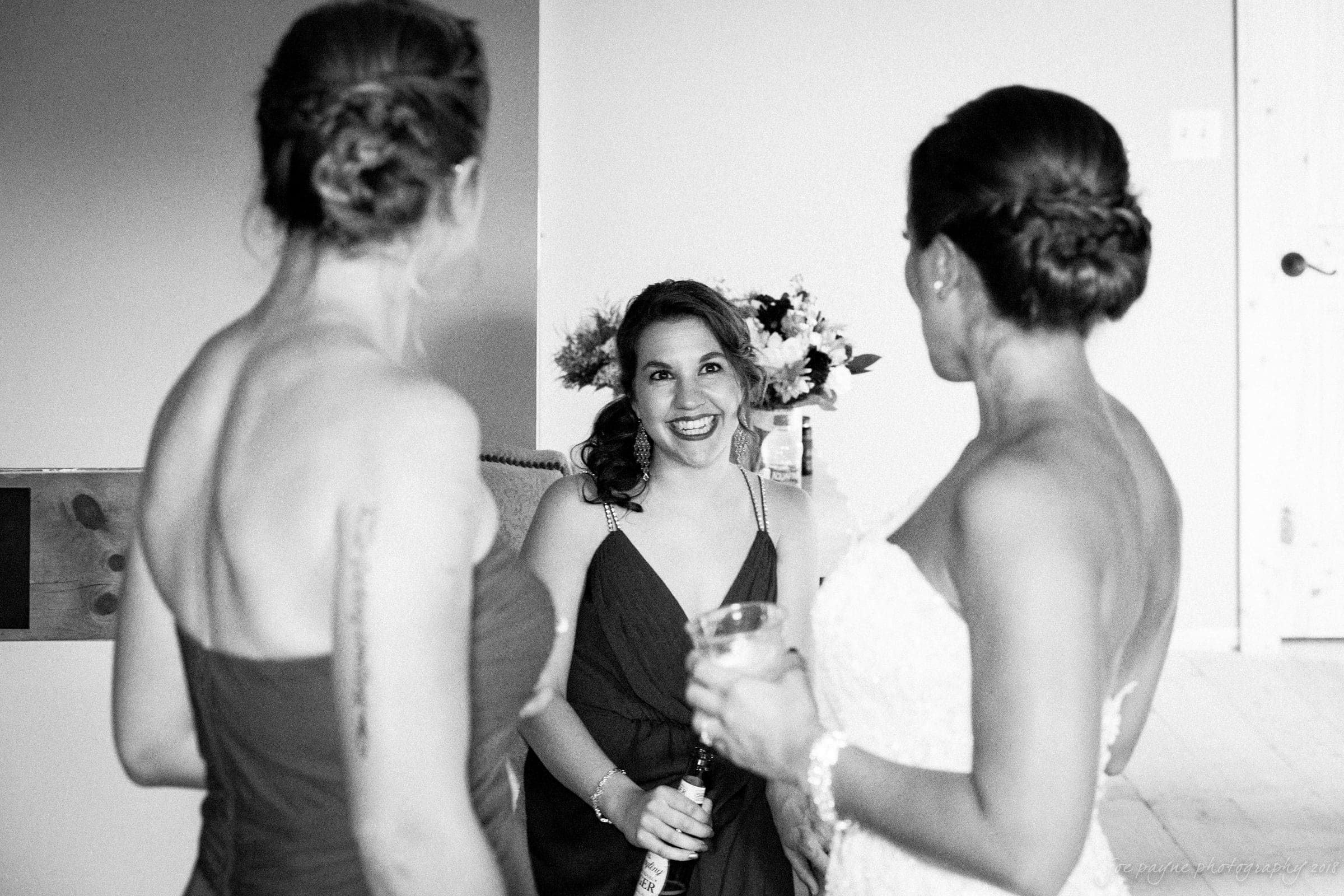pavilion at carriage farm wedding - ashley & devon Three women are indoors, with two standing in the foreground facing away from the camera, and one sitting in the background smiling at them. The scene appears candid and joyful. Black and white photography.