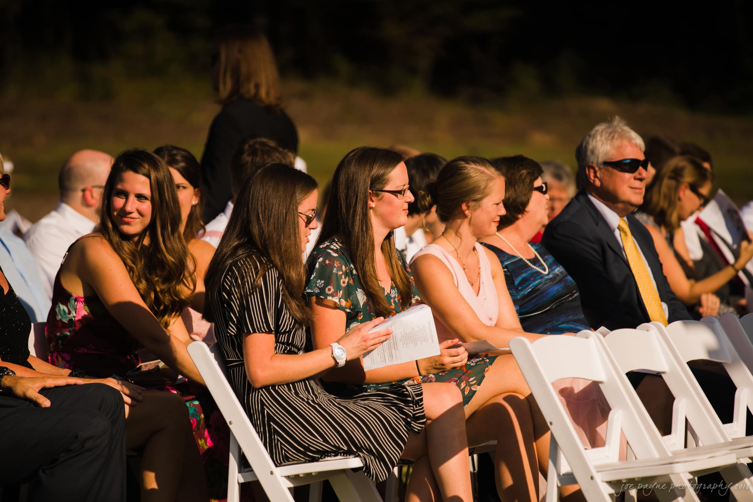 pavilion at carriage farm wedding - ashley & devon A group of people sitting outdoors on white chairs at a formal event. They are dressed in summer clothing, including dresses and suits. Some are holding programs, and the sunlight highlights the scene. The background is blurred vegetation.