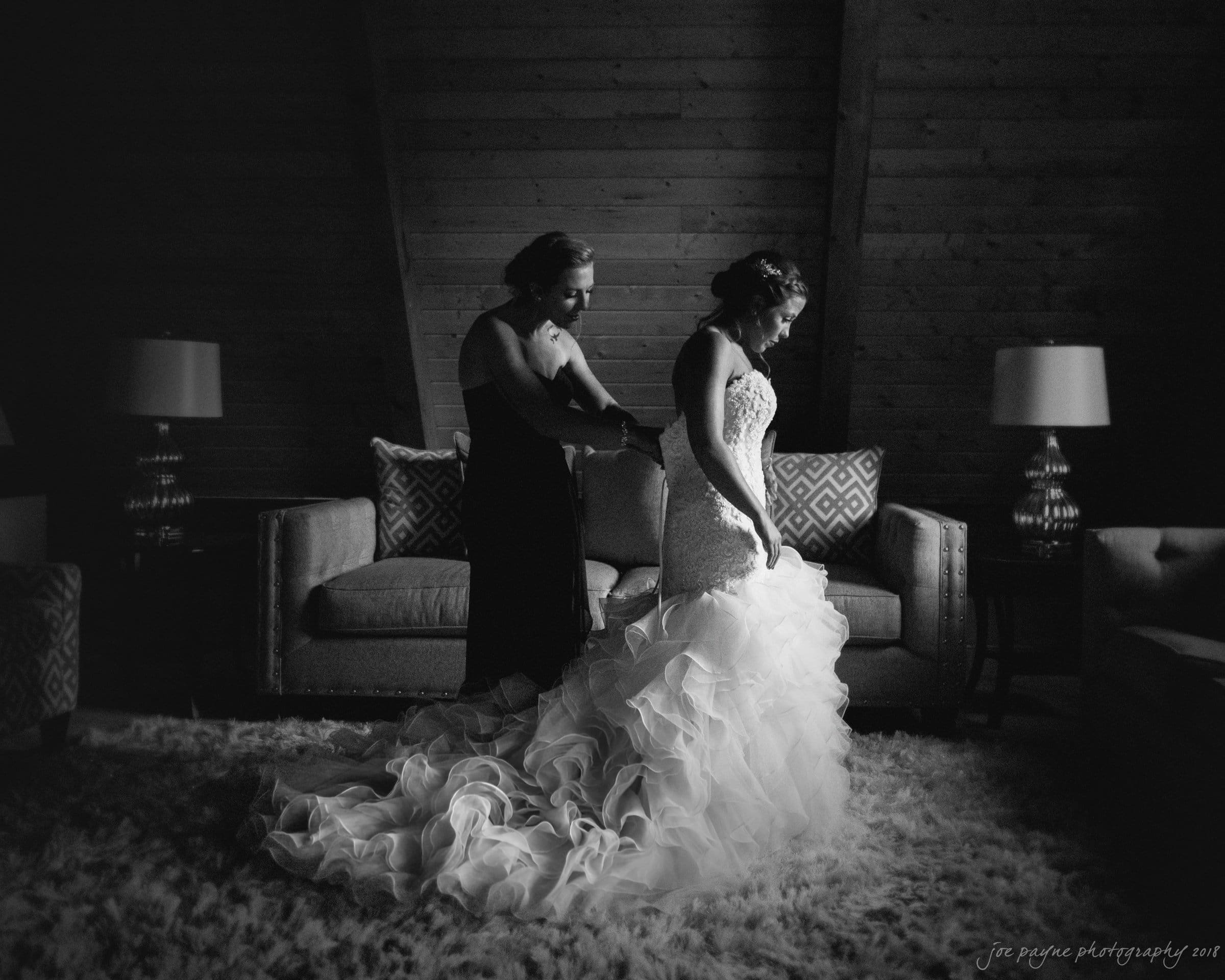 pavilion at carriage farm wedding - ashley & devon A bride stands in a room with wooden walls, wearing a long, ruffled wedding gown. Another woman adjusts her dress. Soft lighting casts gentle shadows, creating a serene and intimate atmosphere. Two lamps and sofas are in the background.