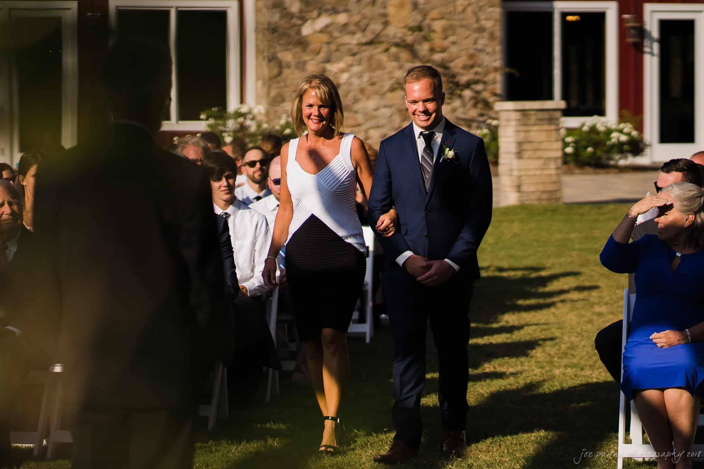 pavilion at carriage farm wedding - ashley & devon A woman and a man, both smiling, walk arm in arm down an outdoor aisle set with white chairs. The audience, seated on either side, watches the pair as they walk on a sunny day. A stone building is visible in the background.
