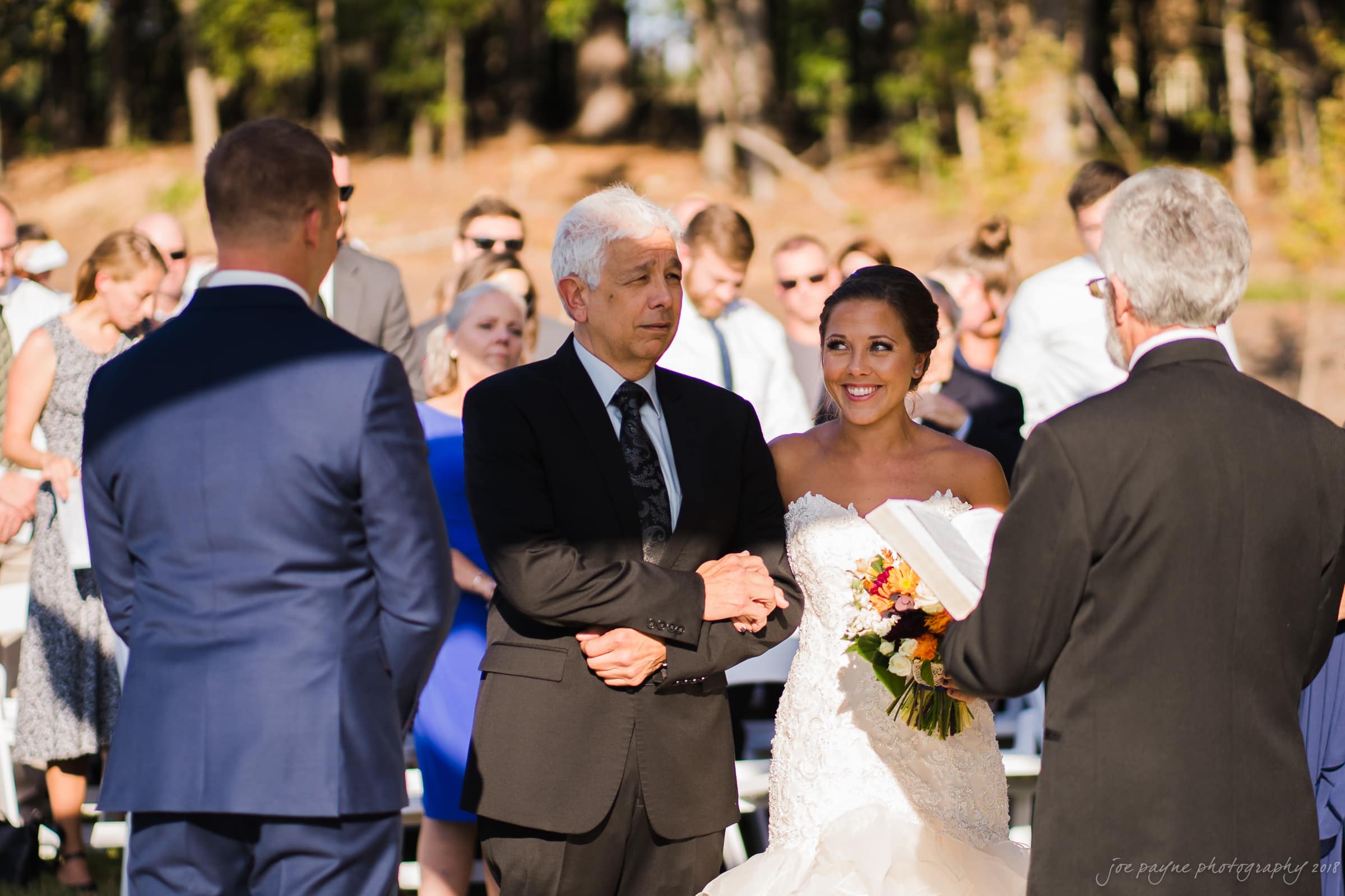 pavilion at carriage farm wedding - ashley & devon A bride in a white dress stands beside an older man in a black suit, holding his arm. The groom, in a blue suit, faces them during an outdoor ceremony. An officiant holds a book, and seated guests watch in the background.