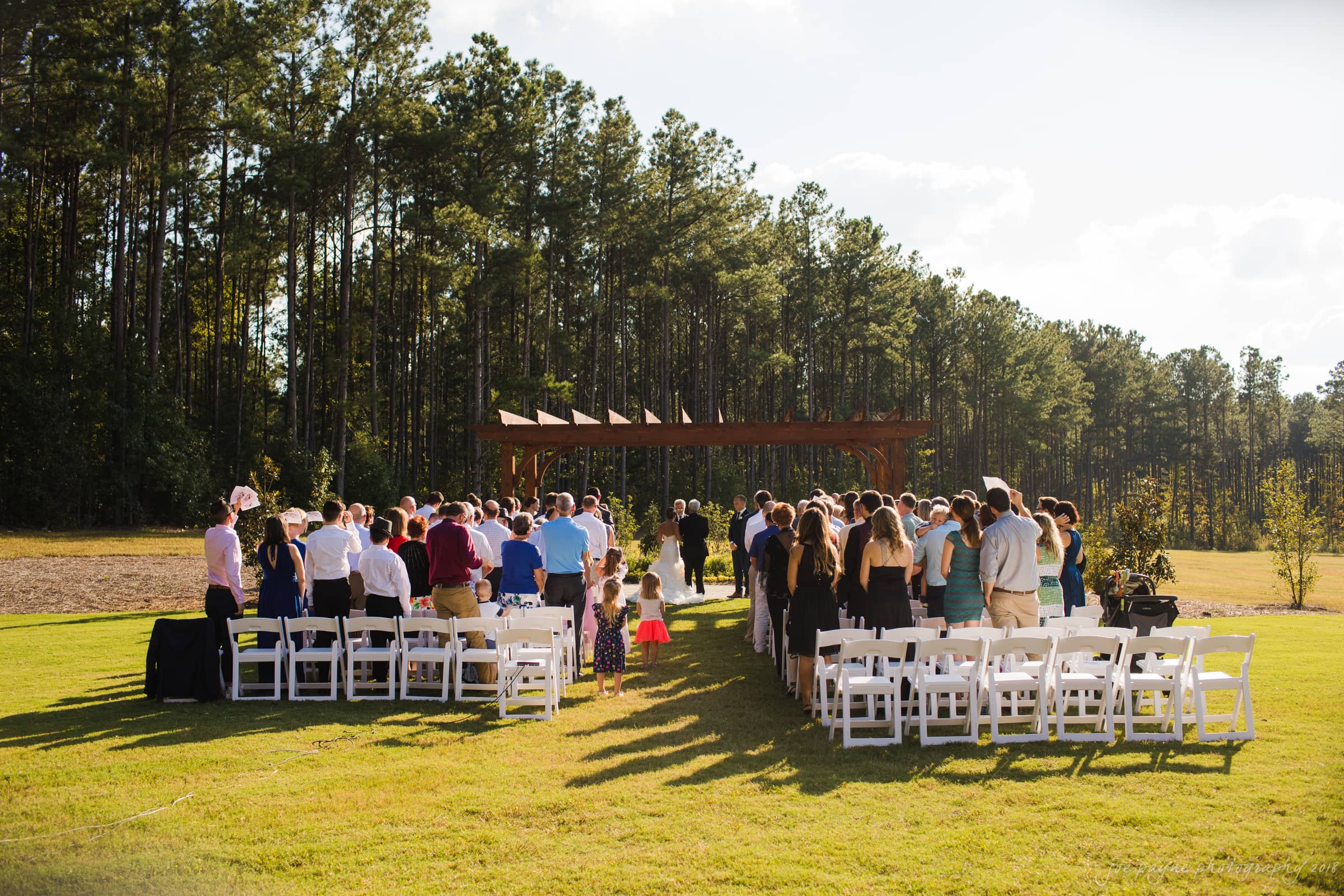 pavilion at carriage farm wedding - ashley & devon A wedding ceremony takes place outdoors on a sunny day. Guests are seated on white chairs facing a wooden arbor, surrounded by tall trees. The bride and groom stand at the altar with a large crowd of attendees watching.