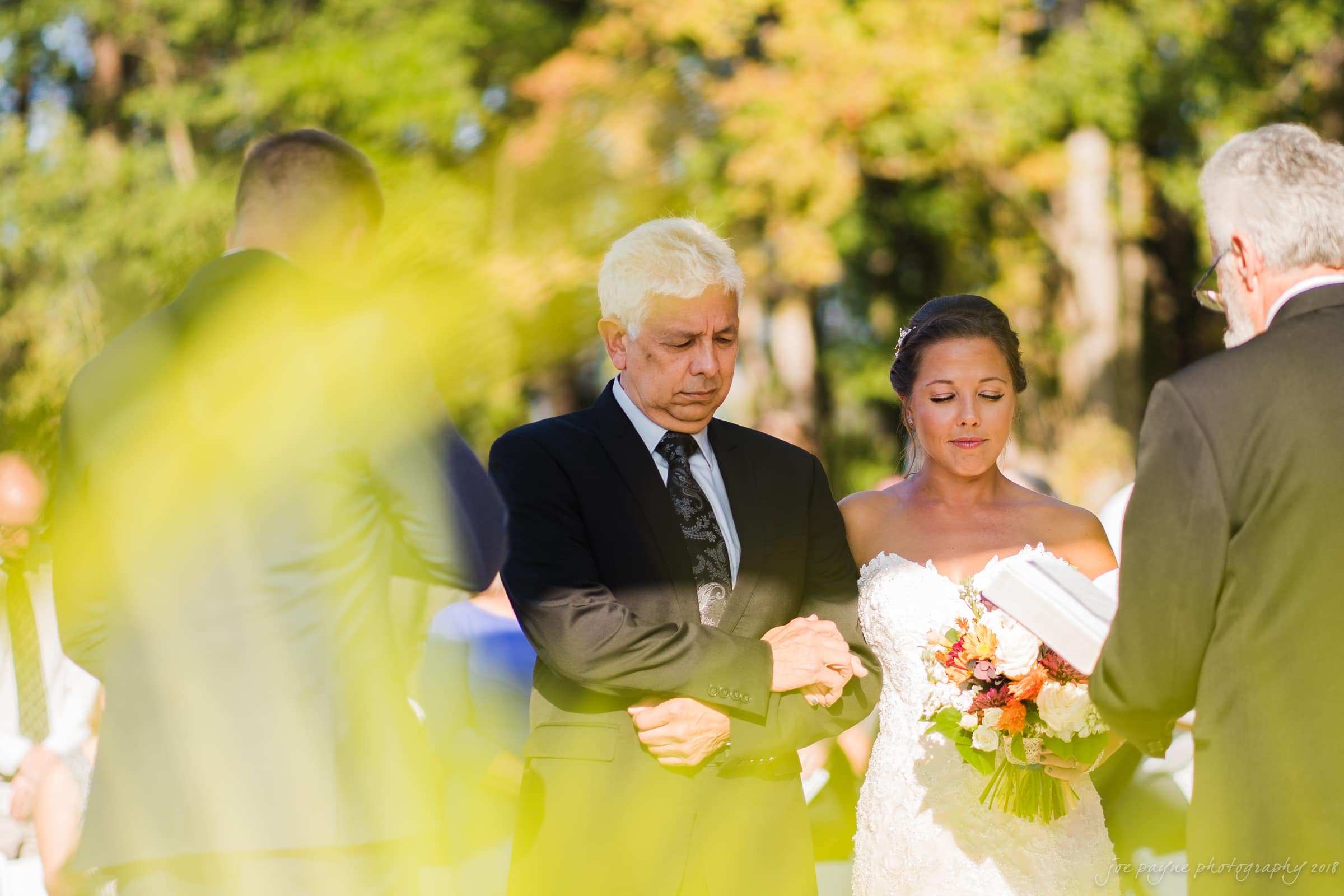 pavilion at carriage farm wedding - ashley & devon A bride and an older man stand outdoors, both with eyes closed, during a wedding ceremony. The bride holds a colorful bouquet, and a person stands in front of them, possibly reading. Sunlight filters through the trees in the background.