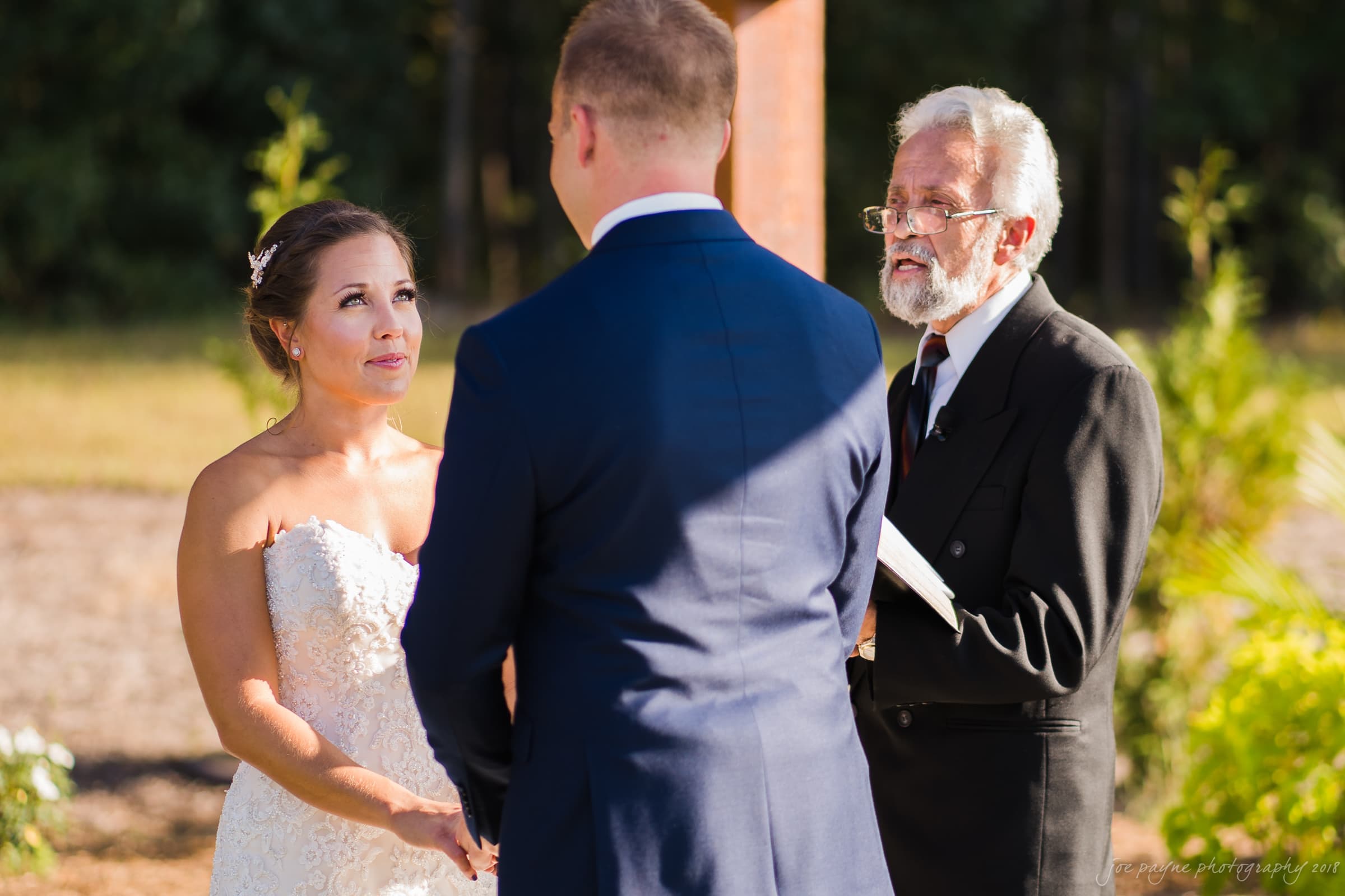 pavilion at carriage farm wedding - ashley & devon A bride in a white dress and a groom in a blue suit stand facing each other, holding hands during an outdoor wedding ceremony. An officiant with a white beard and glasses stands beside them, holding a book. The background is a natural, sunny setting.