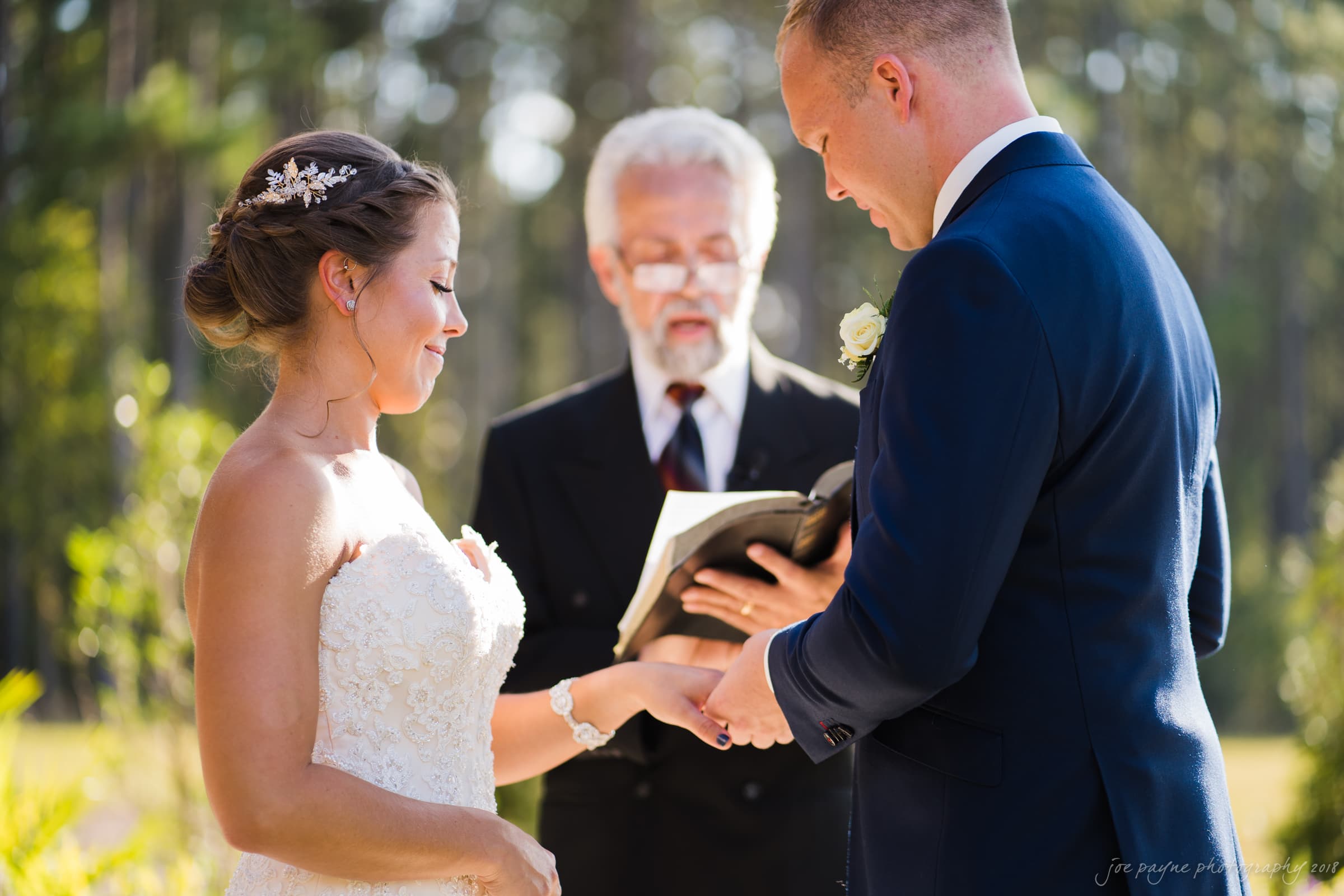 pavilion at carriage farm wedding - ashley & devon A bride and groom stand facing each other, holding hands, during an outdoor wedding ceremony. A man officiating the wedding reads from a book. The background is blurred greenery.