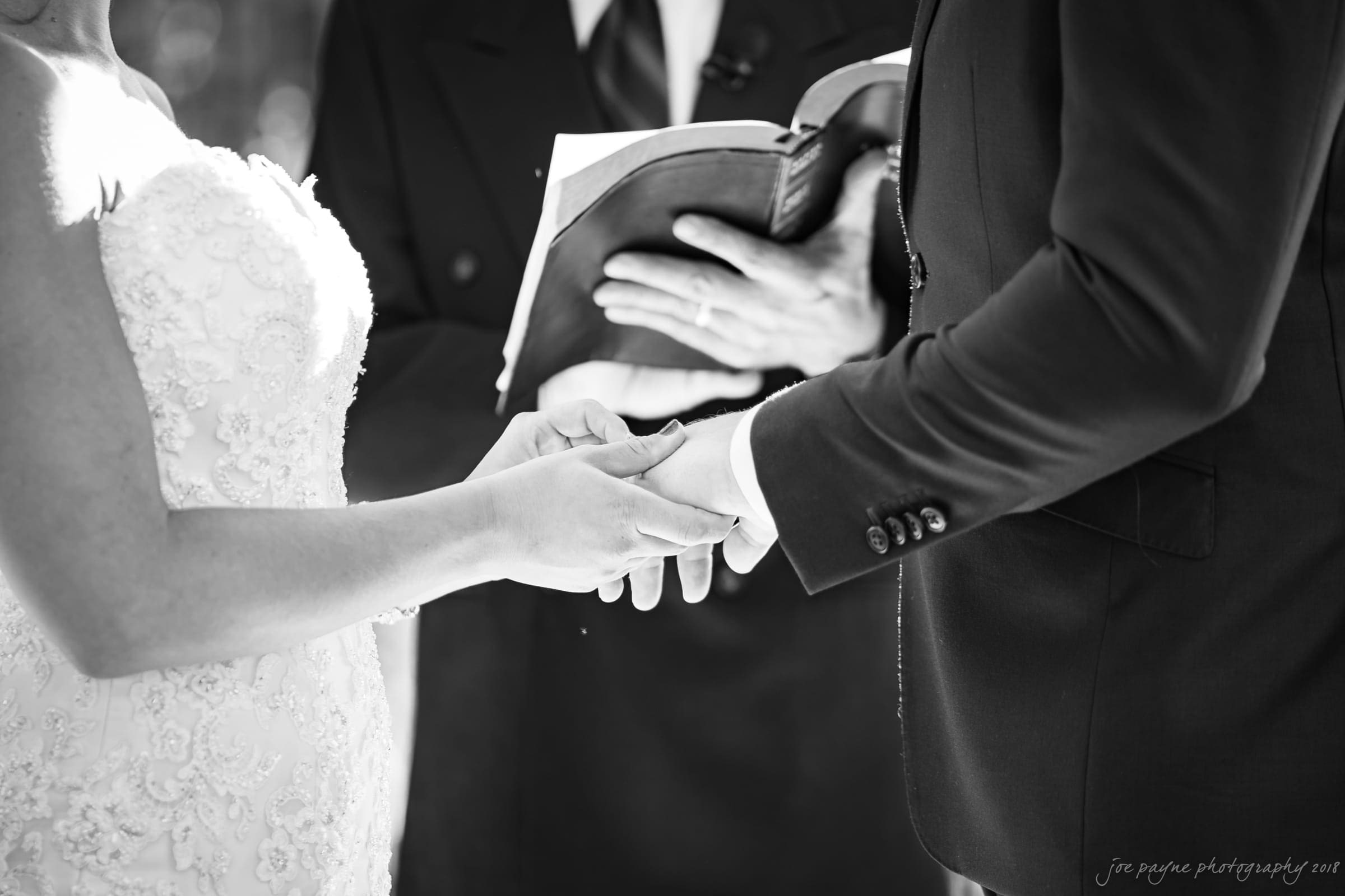 pavilion at carriage farm wedding - ashley & devon A bride and groom exchange vows during a wedding ceremony. They are holding hands, with a person in the background holding an open book, likely officiating the ceremony. The image is in black and white.