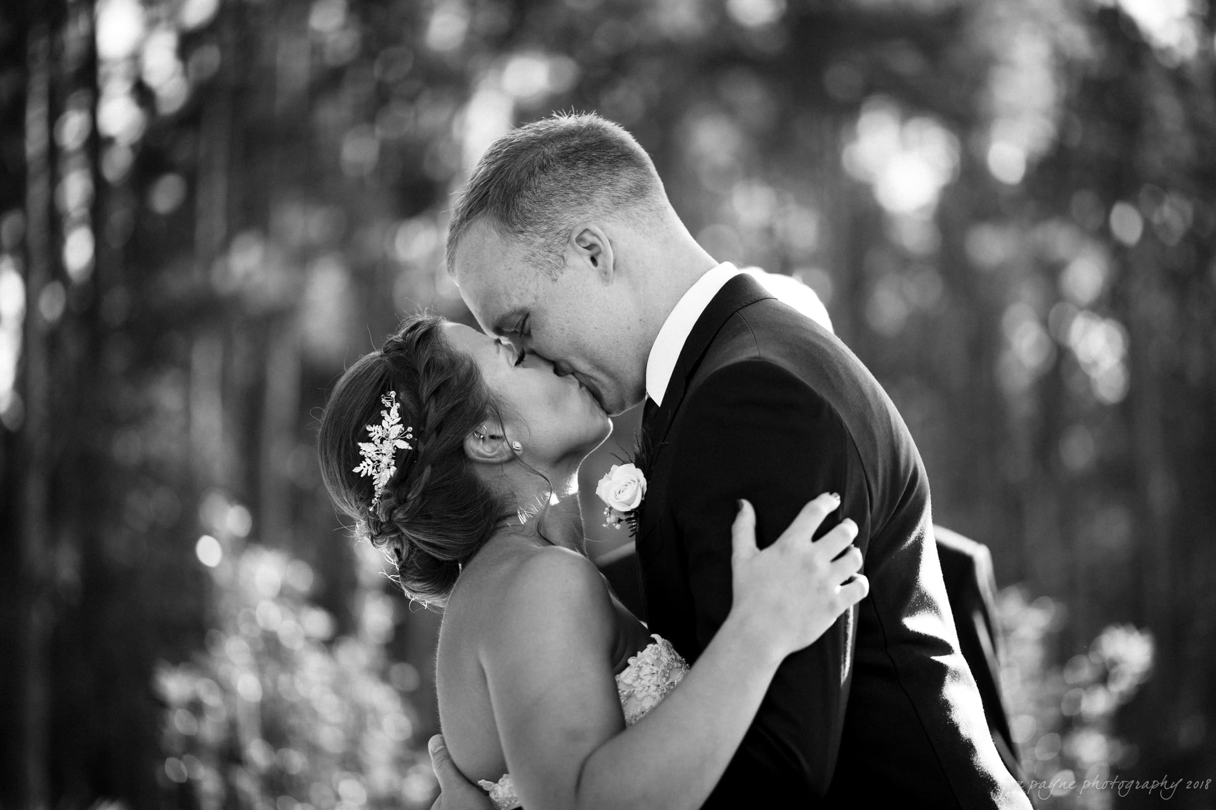pavilion at carriage farm wedding - ashley & devon A bride and groom share a kiss in an outdoor setting. The bride has floral hair accessories and the groom wears a suit with a boutonniere. They embrace each other against a blurred natural background.