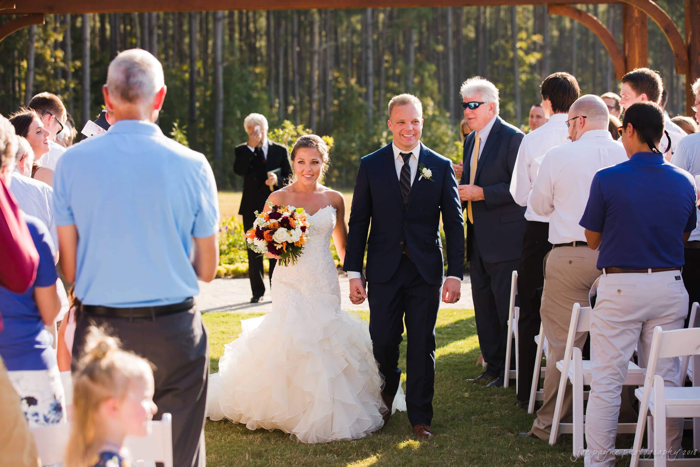 pavilion at carriage farm wedding - ashley & devon A bride and groom in wedding attire walk hand in hand down an outdoor aisle. The bride holds a bouquet of autumn flowers. Guests stand and smile as the couple passes, under a wooden arch, surrounded by trees.