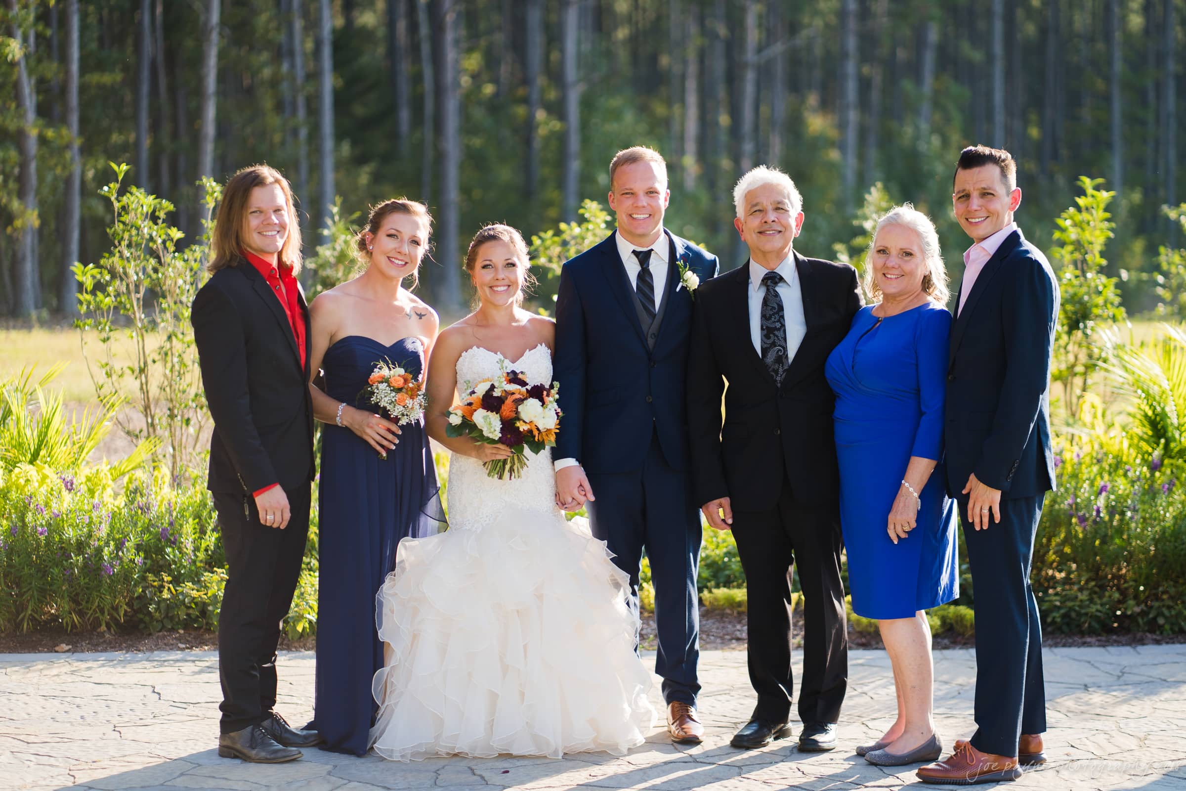 pavilion at carriage farm wedding - ashley & devon A group photo at an outdoor wedding. The bride in a white gown and the groom in a navy suit stand in the center, flanked by family and friends in formal attire. They are all smiling, with trees and greenery in the background.