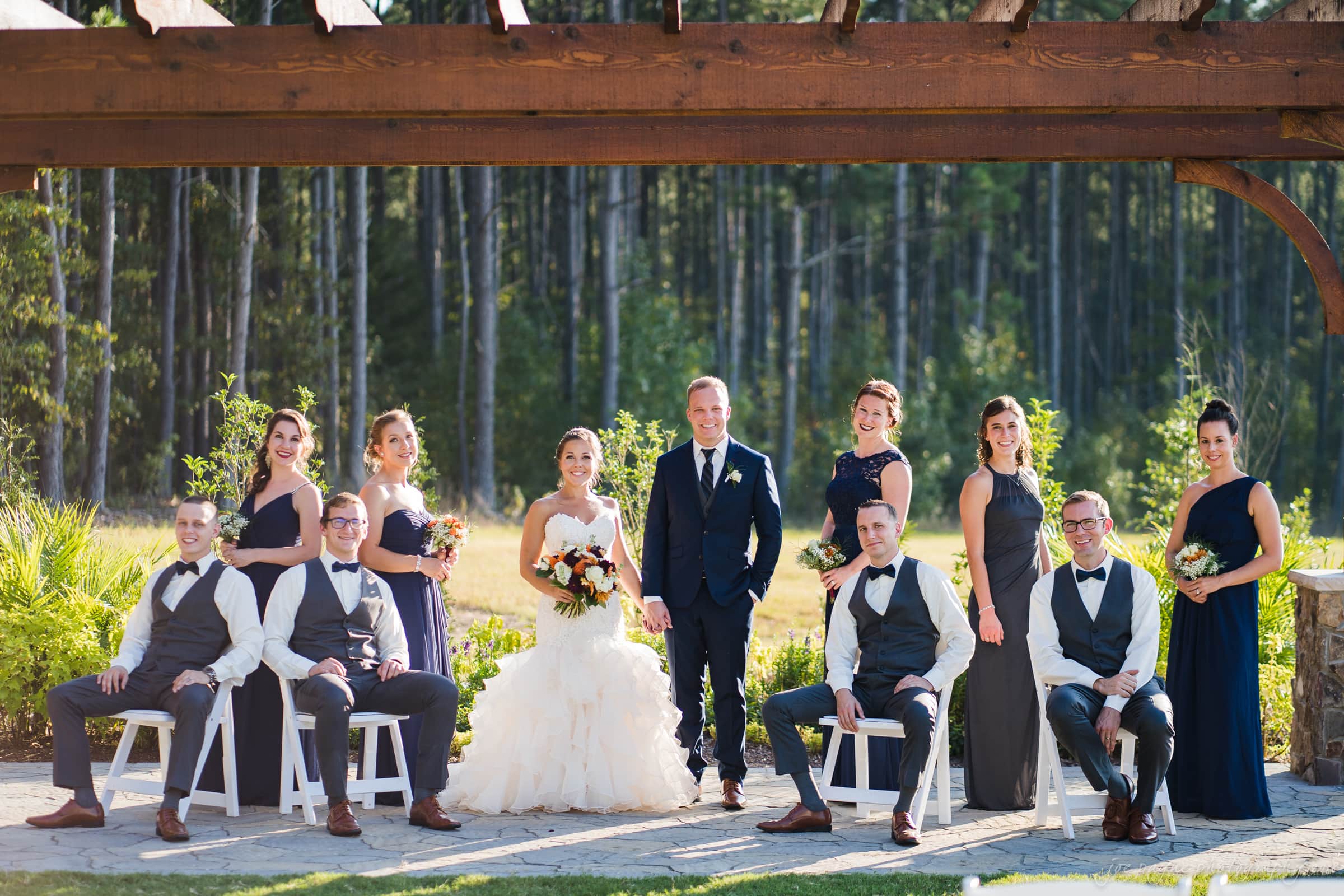 pavilion at carriage farm wedding - ashley & devon A wedding party poses under a wooden pergola in a garden. The bride is in a white gown, the groom in a navy suit. Bridesmaids in navy dresses and groomsmen in vests and bow ties stand and sit around them. Trees can be seen in the background.