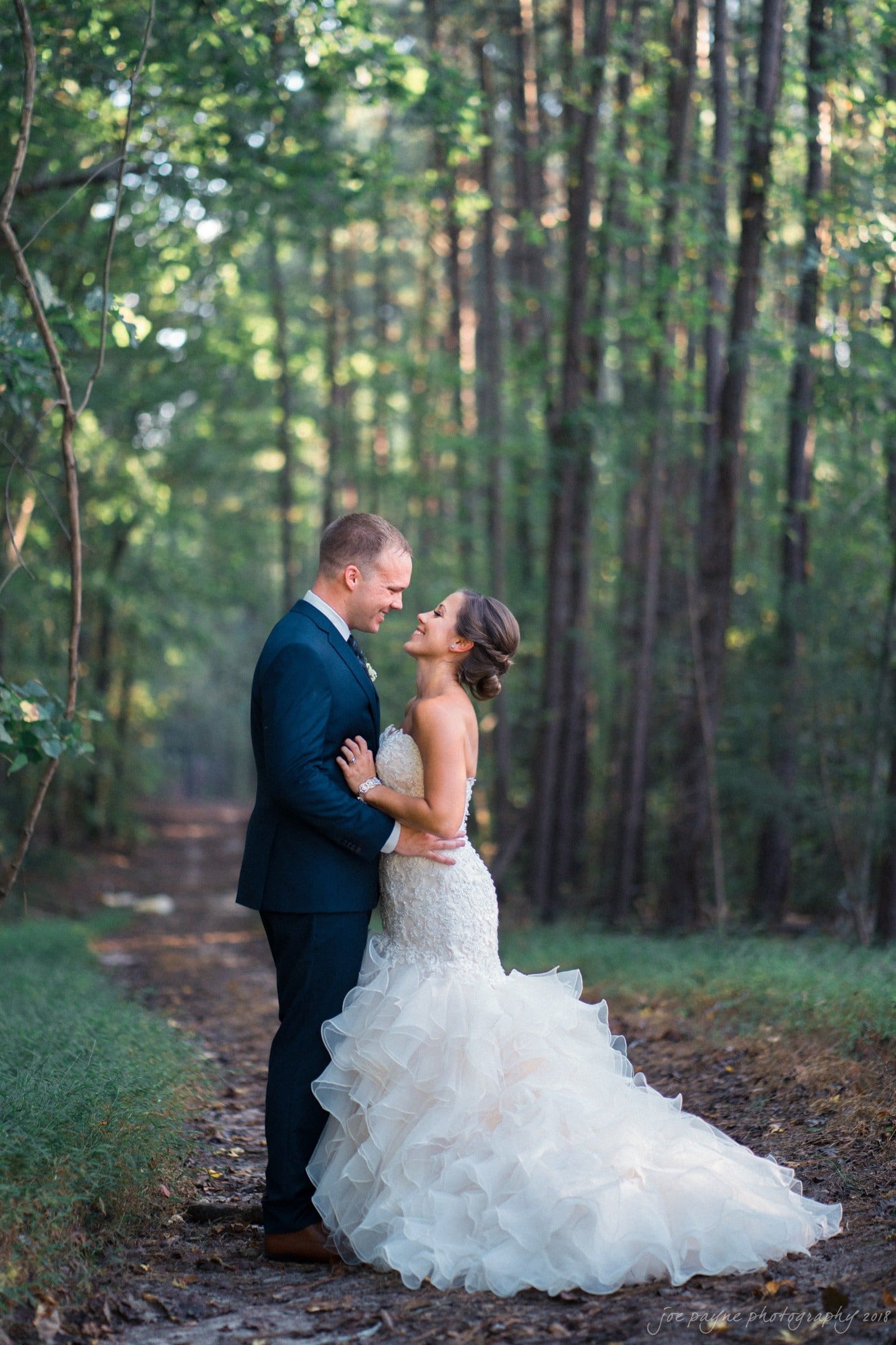 pavilion at carriage farm wedding - ashley & devon A couple stands in a forest path, sharing an intimate moment. The woman wears a flowing, strapless white gown, and the man is in a dark suit. Sunlight filters through the tall trees, casting a soft glow around them.