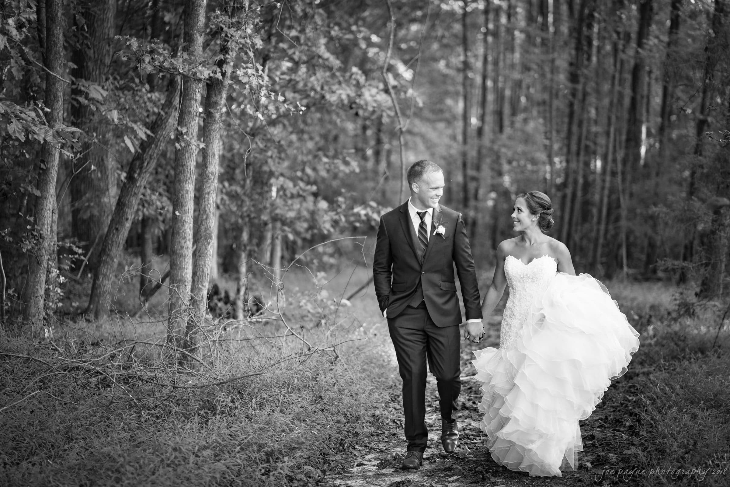 pavilion at carriage farm wedding - ashley & devon A bride and groom walk hand in hand along a forest path. The groom is in a dark suit, and the bride wears a flowing white gown. They are smiling at each other, surrounded by tall trees. The image is in black and white.