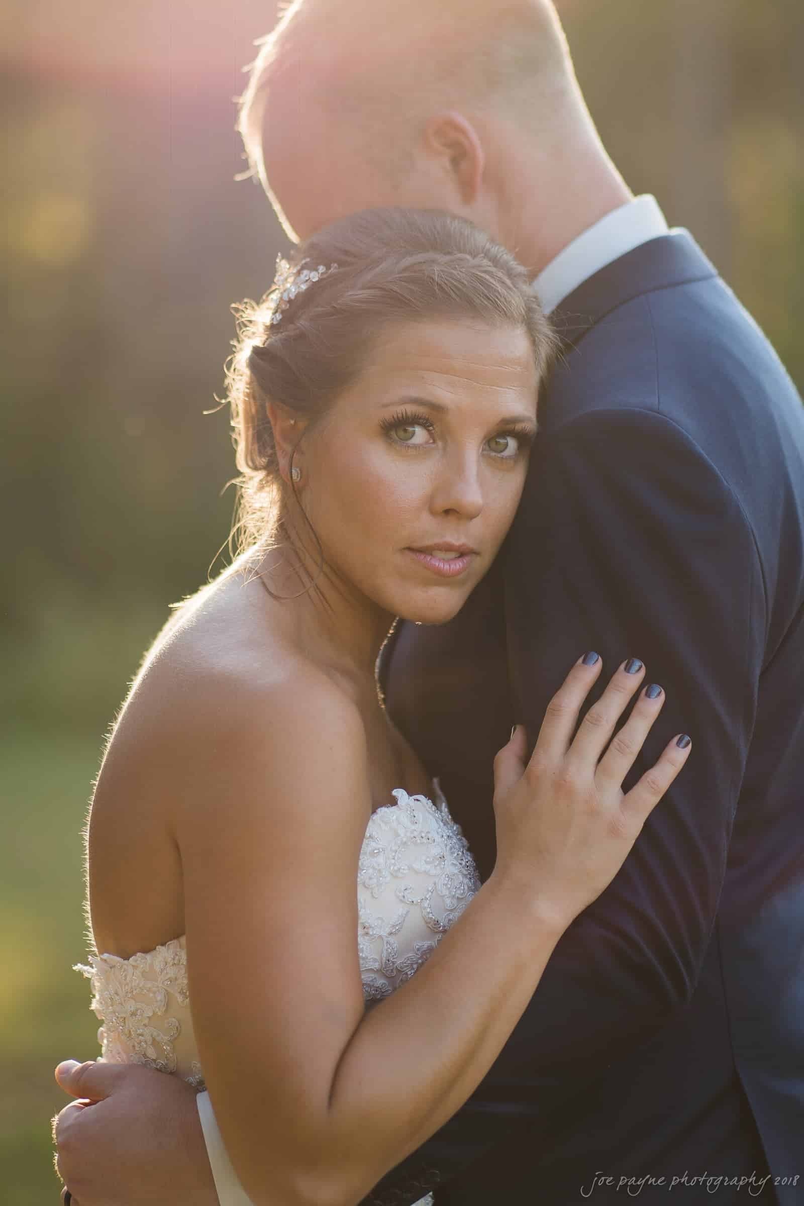 pavilion at carriage farm wedding - ashley & devon A bride in a white dress embraces a groom in a dark suit in a sunlit outdoor setting. The woman looks into the camera with a serious expression, while the mans head is turned away. The sunlight creates a warm, glowing effect.