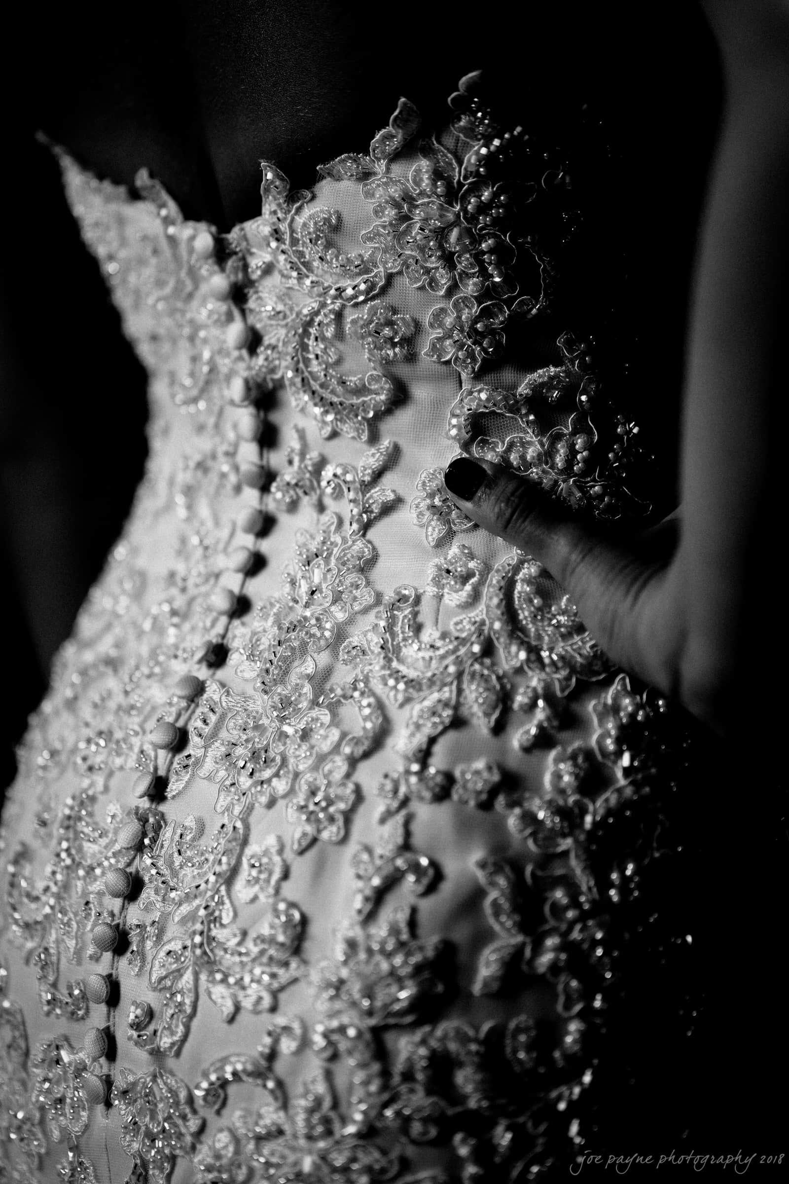 pavilion at carriage farm wedding - ashley & devon Black and white image of a close-up on an intricately detailed bridal gown, featuring floral lace patterns and delicate buttons. A hand with painted nails rests on the hip, enhancing the elegance of the ensemble.