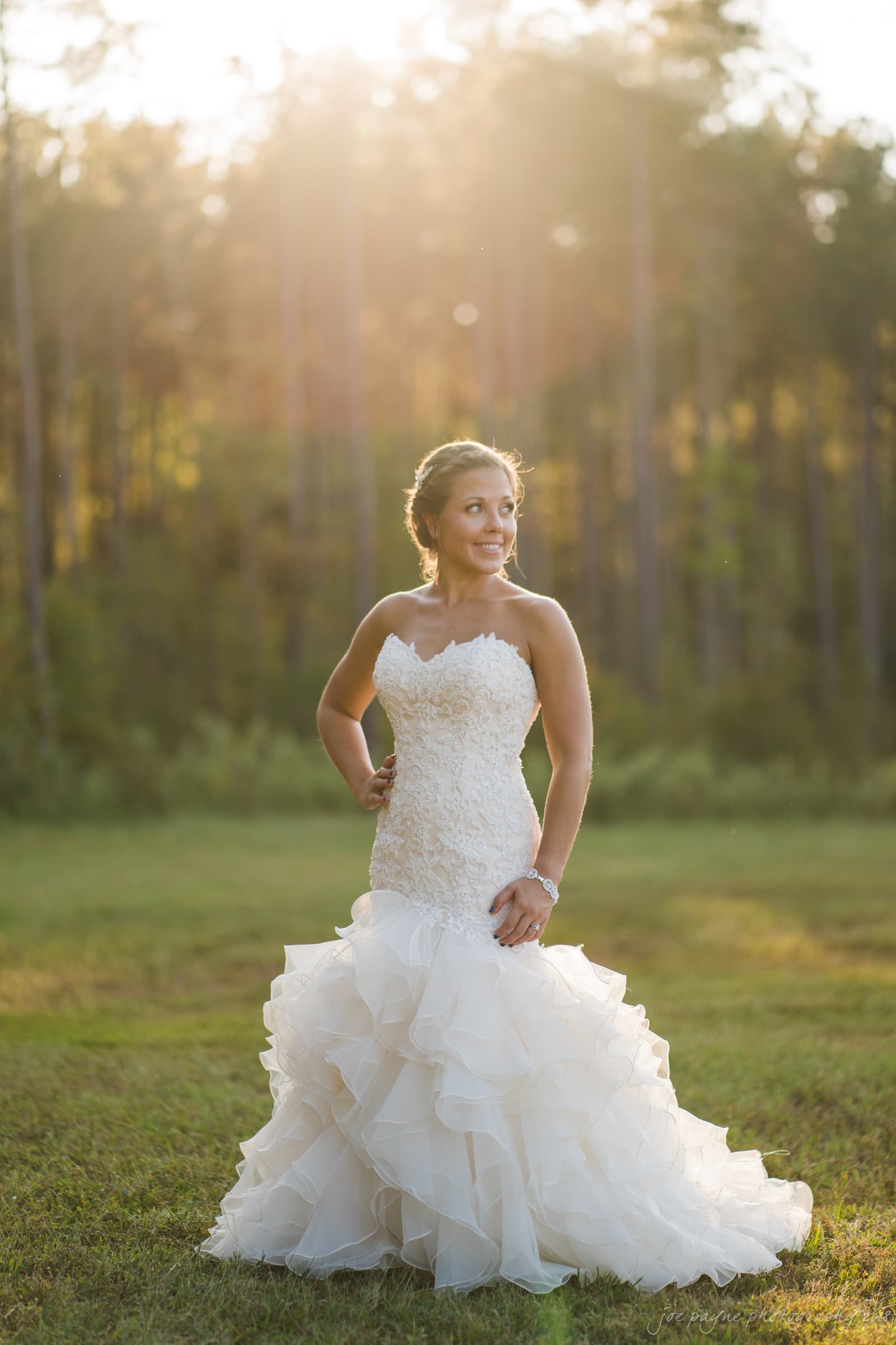 pavilion at carriage farm wedding - ashley & devon A woman in a strapless, ruffled wedding dress stands confidently outdoors. She is smiling, with hands on her hips, in front of a blurred forest background. The sunlight creates a halo effect around her, enhancing the serene atmosphere.