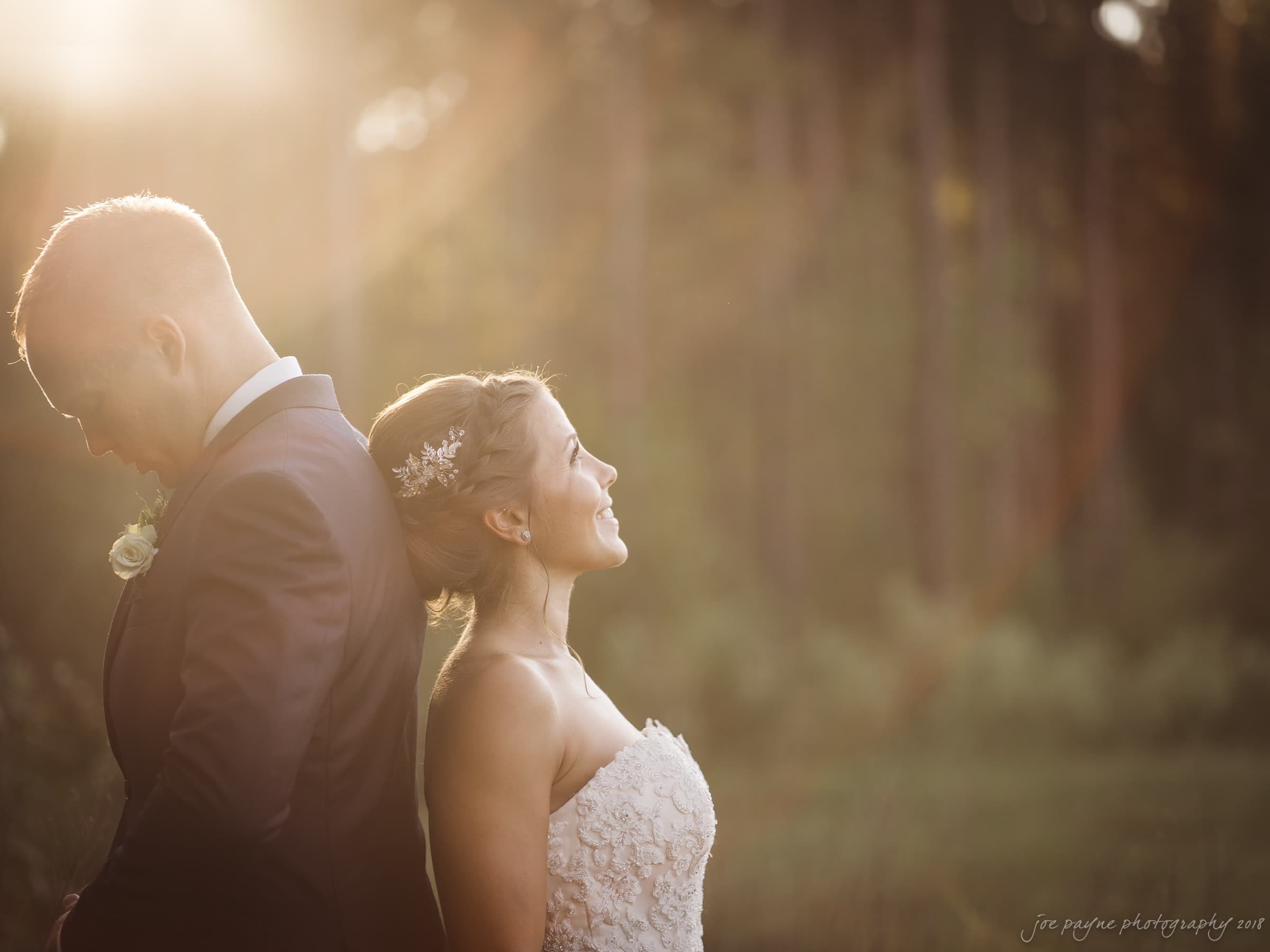 pavilion at carriage farm wedding - ashley & devon A bride and groom stand back-to-back in a forest during sunset. The bride wears a strapless white gown with a floral hair accessory, and the groom wears a dark suit with a white rose boutonniere. Sunlight filters through the trees, creating a warm glow.