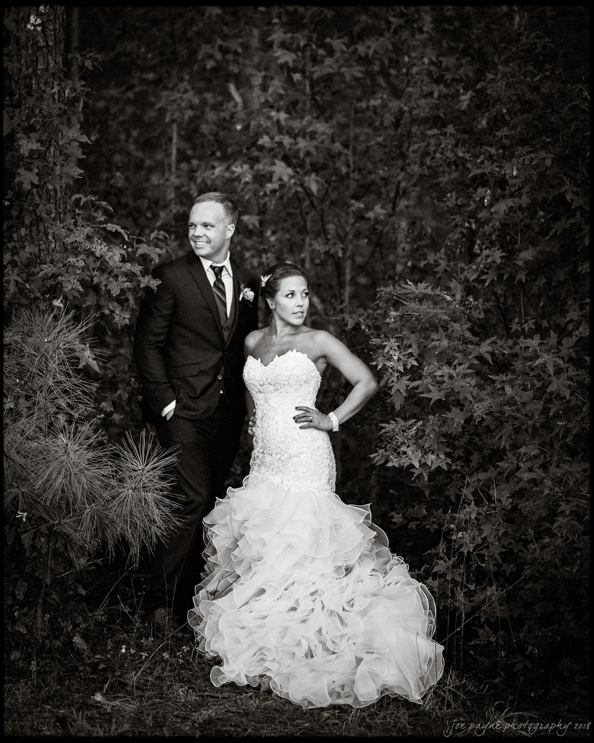 pavilion at carriage farm wedding - ashley & devon A bride in a flowing, strapless wedding gown and a groom in a dark suit stand together in a forested area. The groom looks to the side with his hands in his pockets, while the bride poses with her hand on her hip. The image is in black and white.