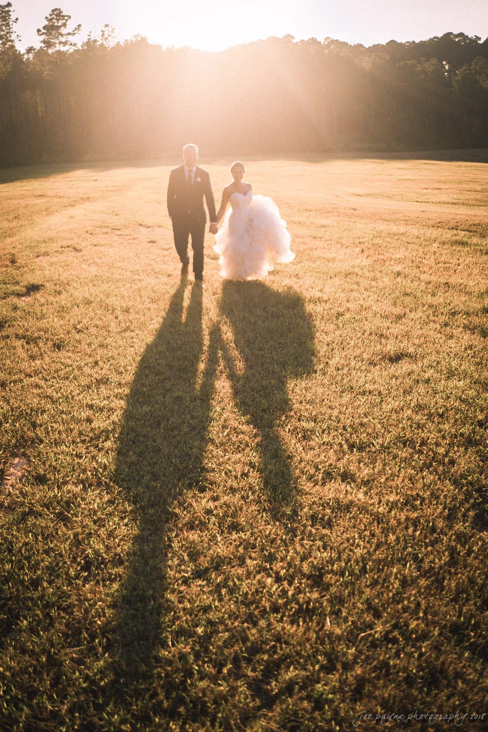 pavilion at carriage farm wedding - ashley & devon A couple, dressed in formal attire, walks hand in hand across a sunlit field. The long shadows cast by the setting sun enhance the warm, romantic atmosphere. The womans dress cascades in soft layers as they stroll toward the horizon.