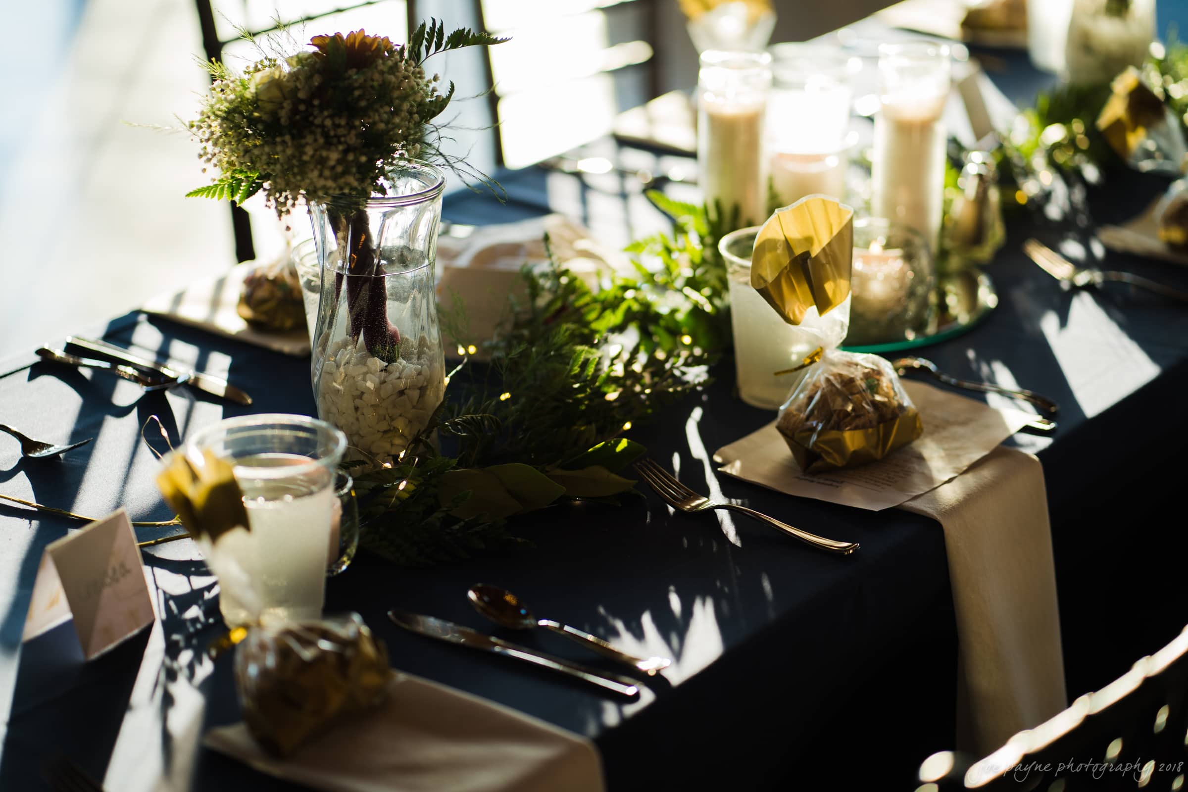 pavilion at carriage farm wedding - ashley & devon A sunlit table set for an event, featuring elegant decorations. There are white napkins, wrapped gifts, glass vases with pebbles and greenery, and candles on the dark tablecloth, creating a warm and inviting atmosphere.