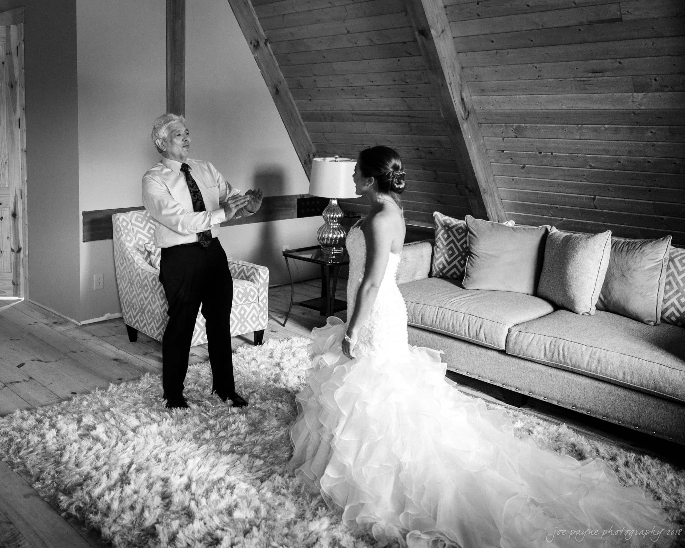 pavilion at carriage farm wedding - ashley & devon A black and white photo of a bride in a ruffled gown stands in a cozy room with wooden ceilings as she shares a joyful moment with an older man in a tie. They are both smiling, surrounded by a plush rug, a sofa, and a lamp.