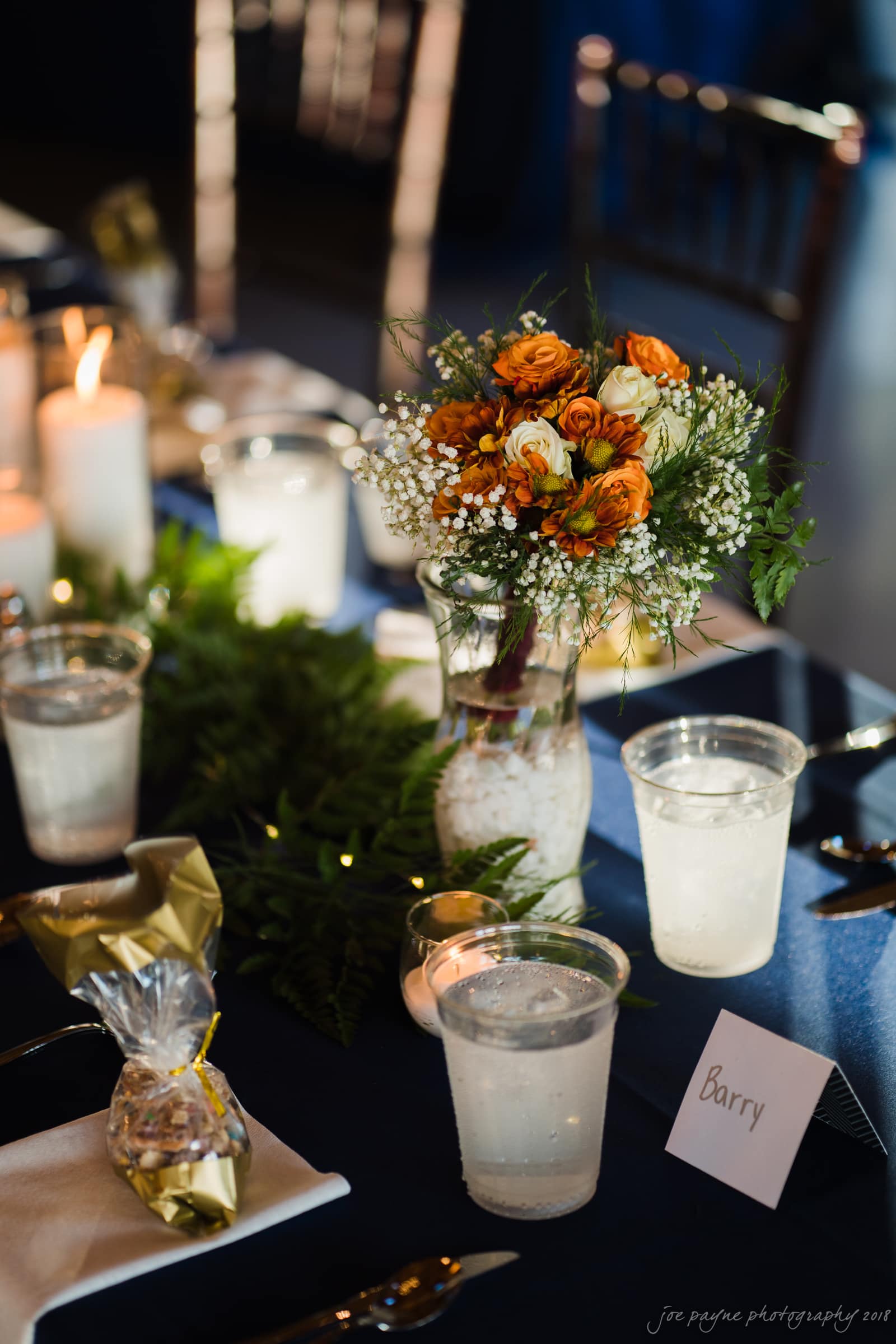 pavilion at carriage farm wedding - ashley & devon A table setting with a floral centerpiece featuring orange and white flowers in a vase. There are candles, water glasses, and a small wrapped gift. A place card labeled Barry is visible on the table.