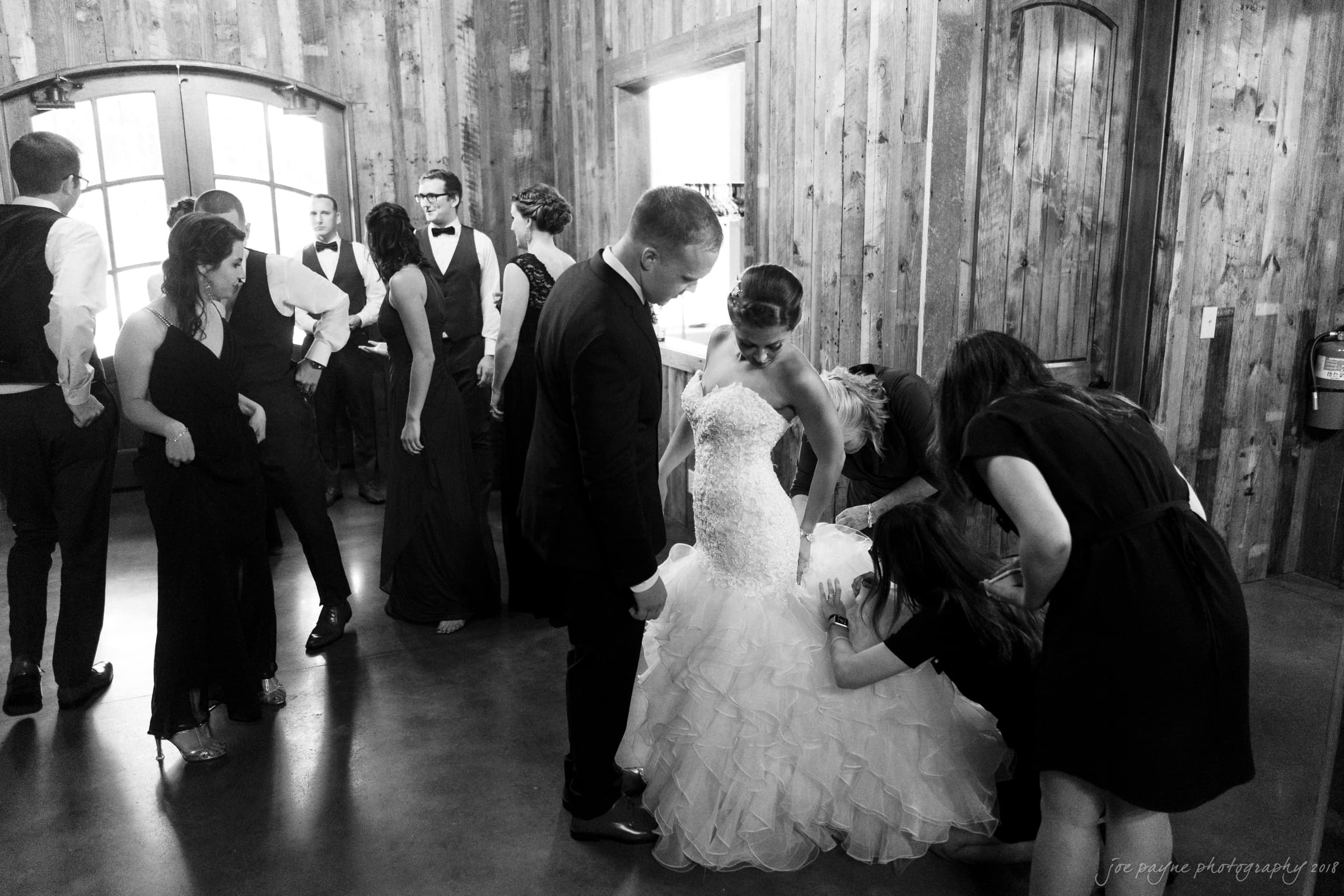pavilion at carriage farm wedding - ashley & devon A bride in a wedding gown is being assisted by several people in a rustic wooden room. Other guests in formal attire are gathered nearby, some chatting and smiling. The atmosphere is lively and festive.