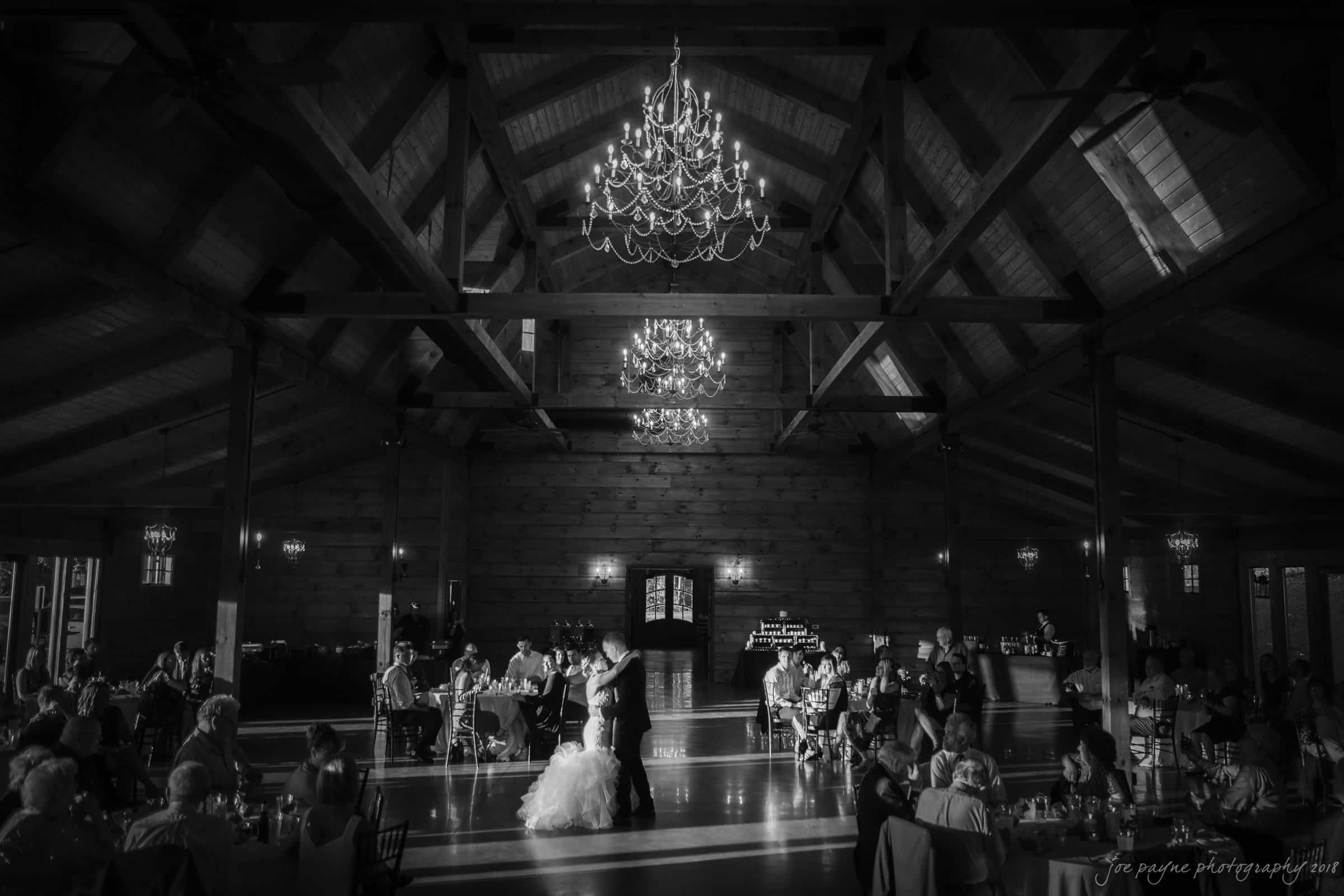 pavilion at carriage farm wedding - ashley & devon A black and white photo captures a wedding reception in a spacious hall with wooden beams. A couple dances under chandeliers, surrounded by seated guests.