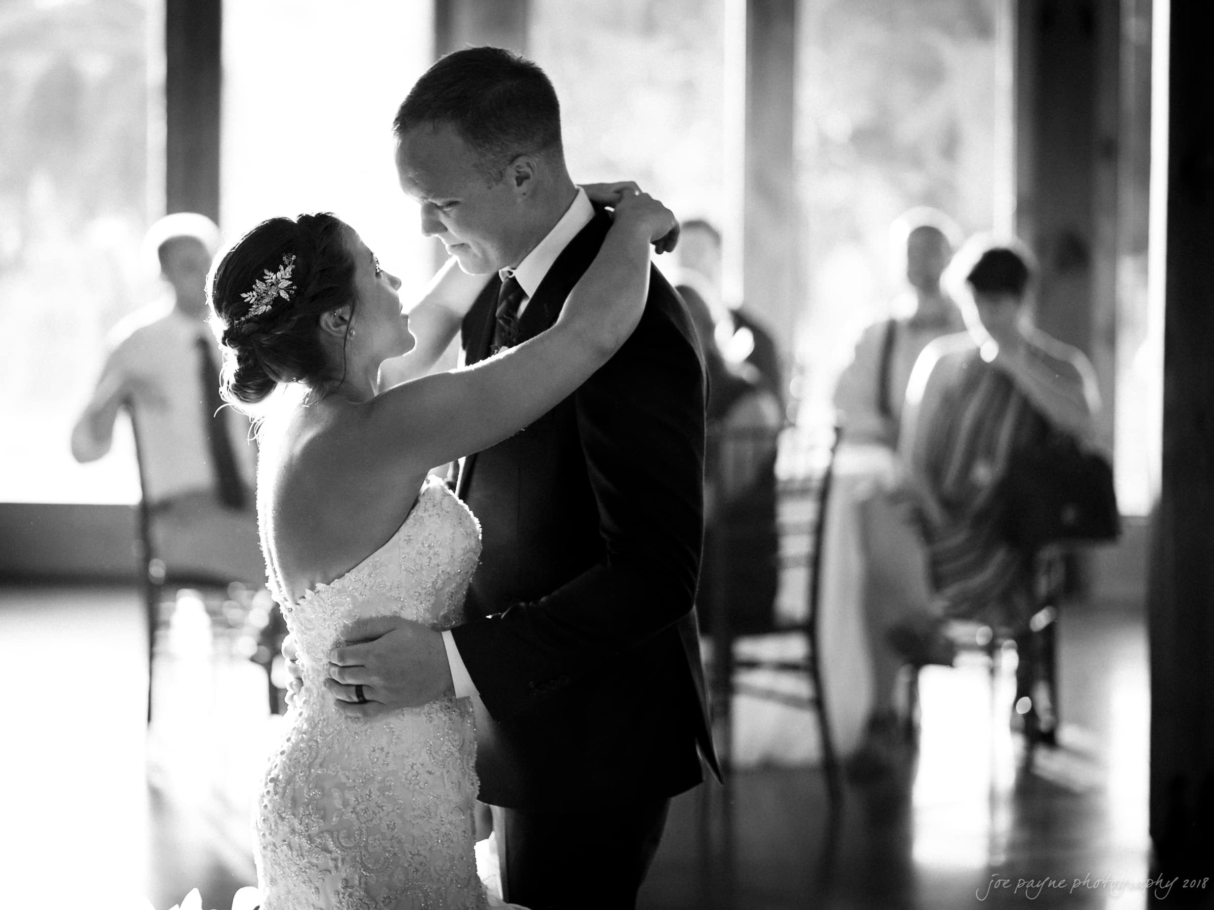 pavilion at carriage farm wedding - ashley & devon A bride and groom share their first dance, smiling at each other in a softly lit room. Guests sit in the background, watching the couple lovingly. The atmosphere is intimate and joyful.