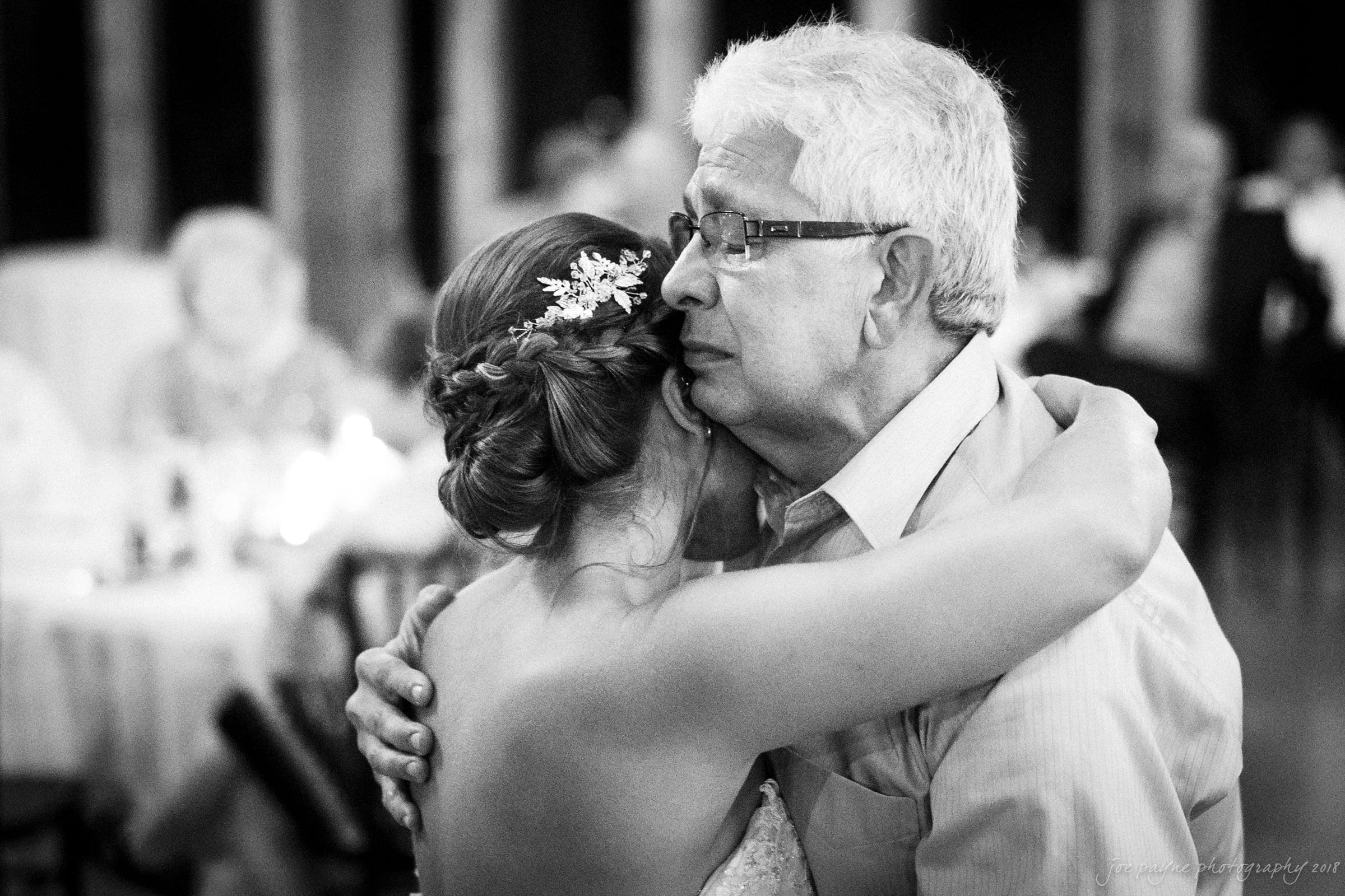 pavilion at carriage farm wedding - ashley & devon A black and white photo of an elderly man and a young woman embracing closely, with the woman’s hair in a braided updo adorned with small flowers. They appear to be dancing in a softly lit room, surrounded by blurred seated guests.