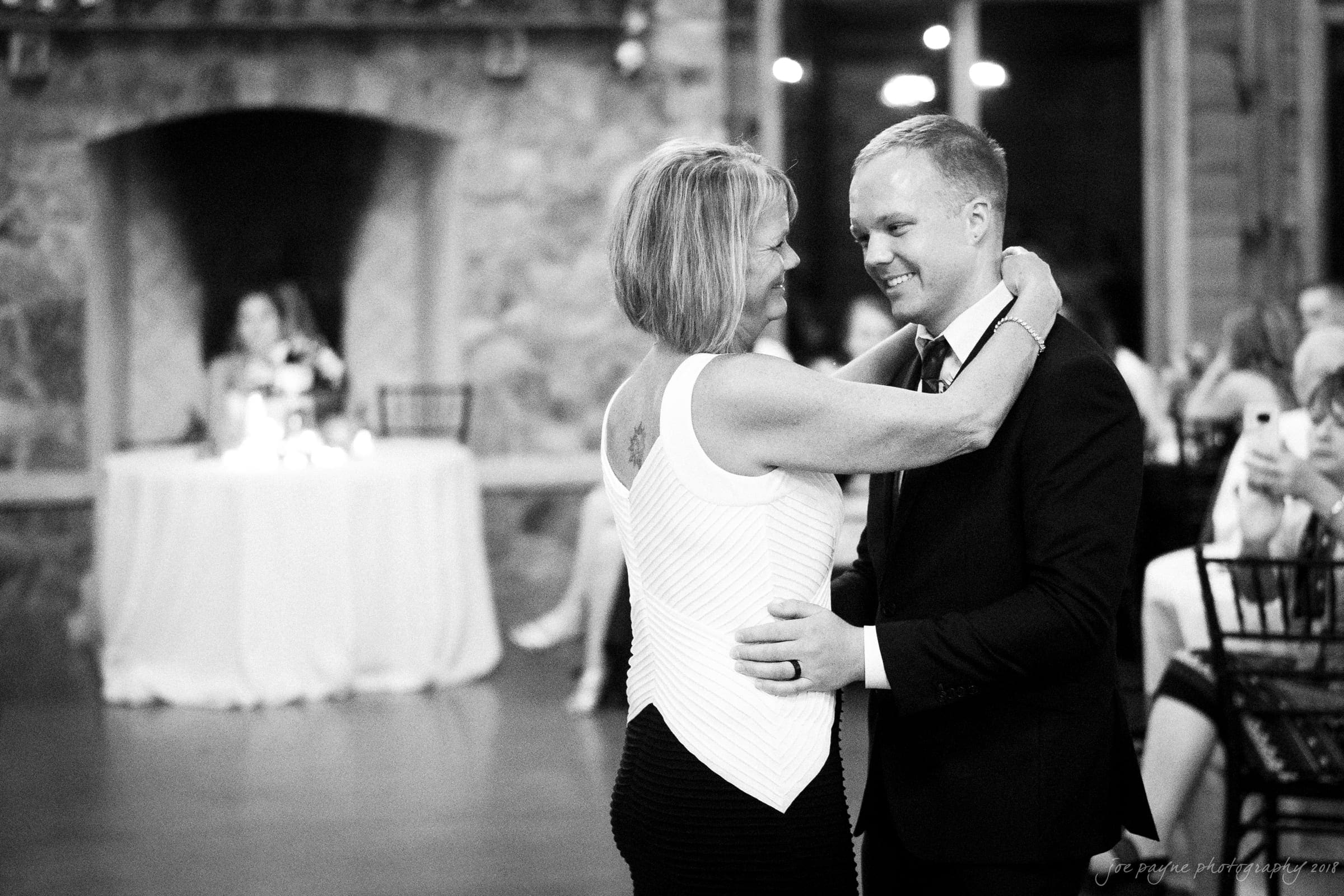 pavilion at carriage farm wedding - ashley & devon A woman and man dance closely, smiling at each other in a warmly lit room with a stone fireplace in the background. People are seated at tables nearby, watching the dance. The mood is joyful and intimate.
