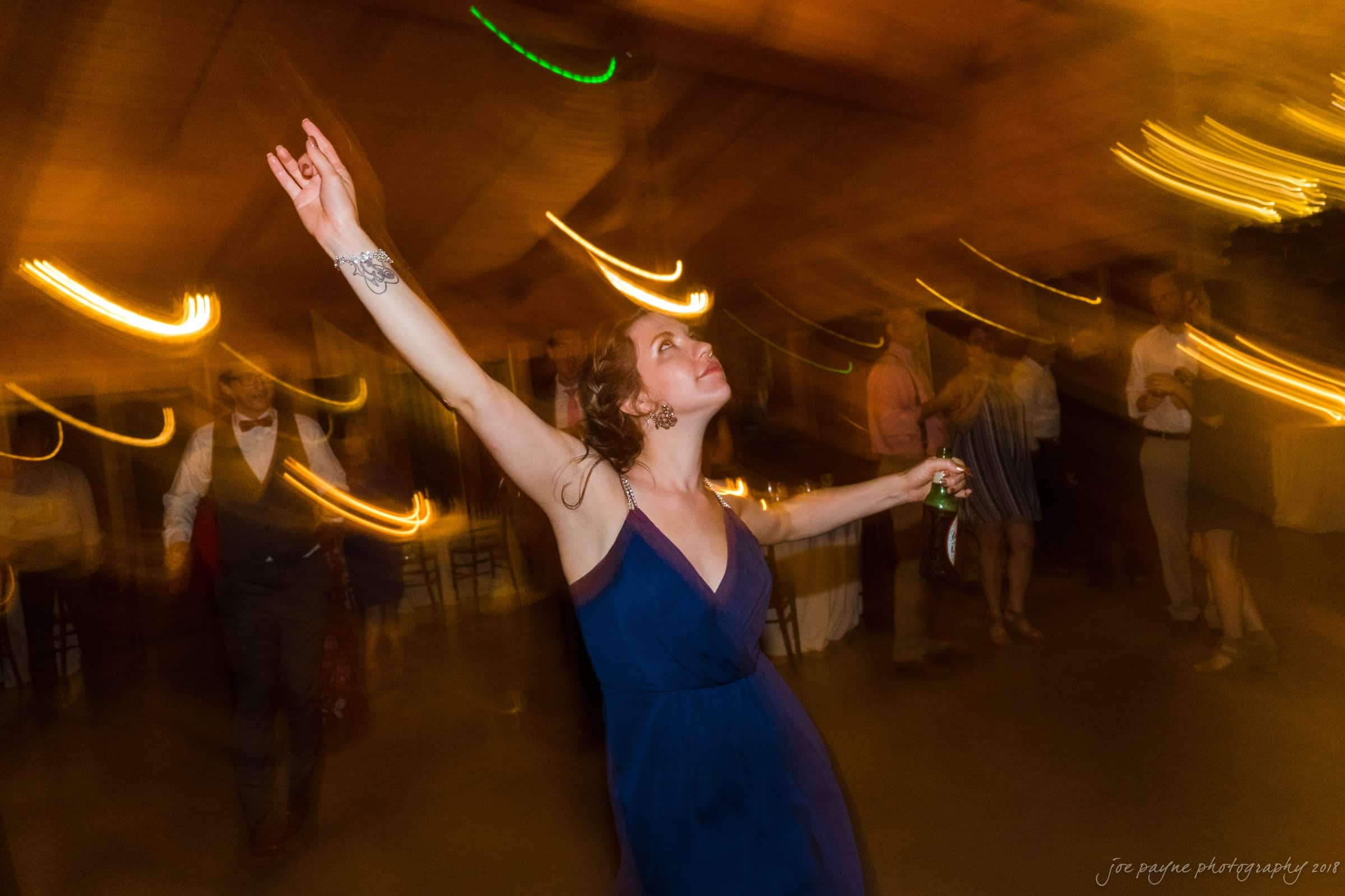 pavilion at carriage farm wedding - ashley & devon A woman in a navy dress dances energetically with her arms raised at a dimly lit event. The background is blurred, capturing motion and colorful lights, creating an energetic and lively atmosphere.