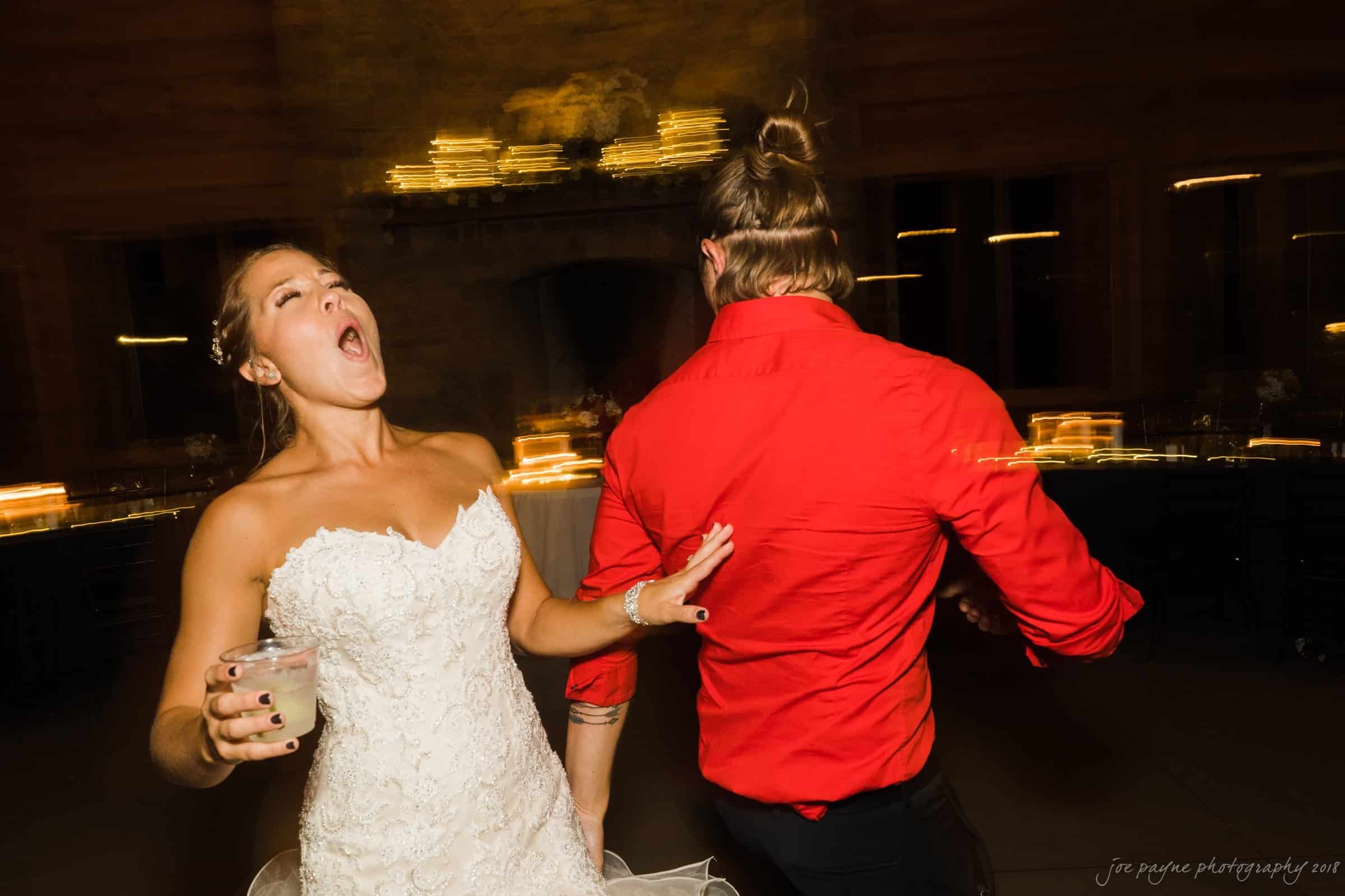 pavilion at carriage farm wedding - ashley & devon A bride in a white dress energetically sings or shouts while dancing with a person in a red shirt. The background is dimly lit with string lights, creating a lively and festive atmosphere.