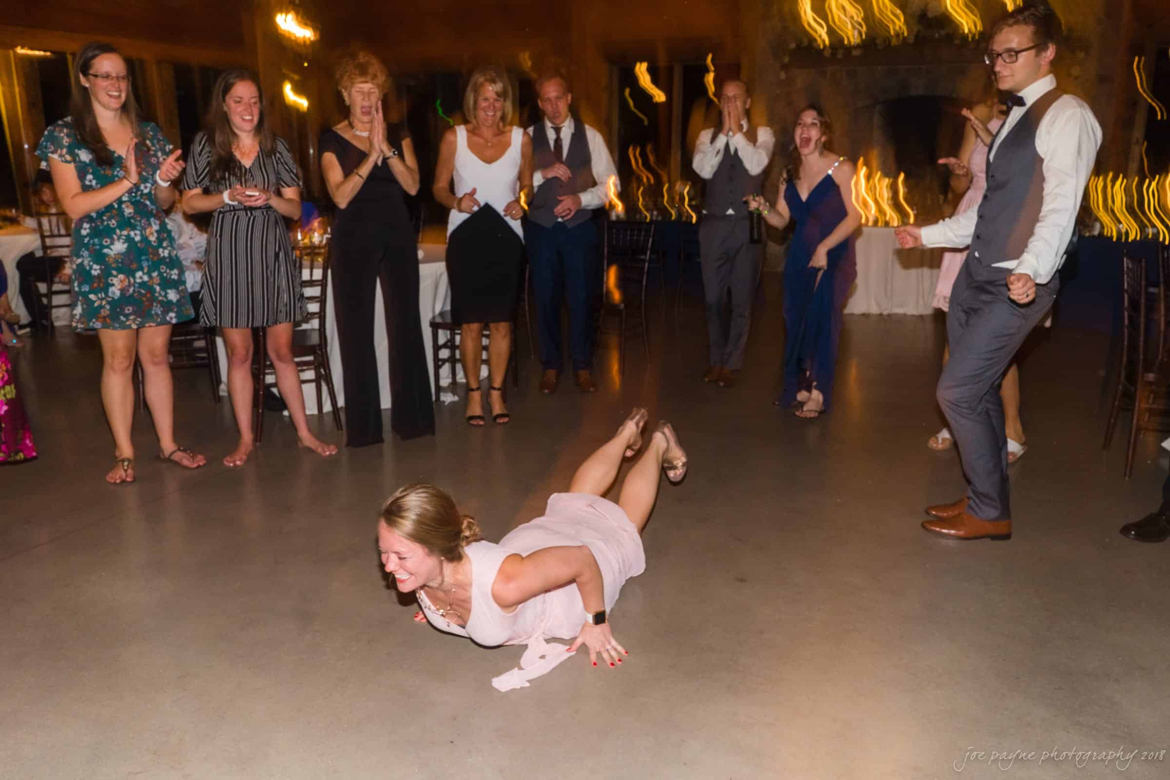 pavilion at carriage farm wedding - ashley & devon A woman in a pink dress is doing the worm dance move on a dance floor, while a group of people surrounds her, smiling and clapping. The background shows blurred lights, indicating movement and a festive atmosphere.