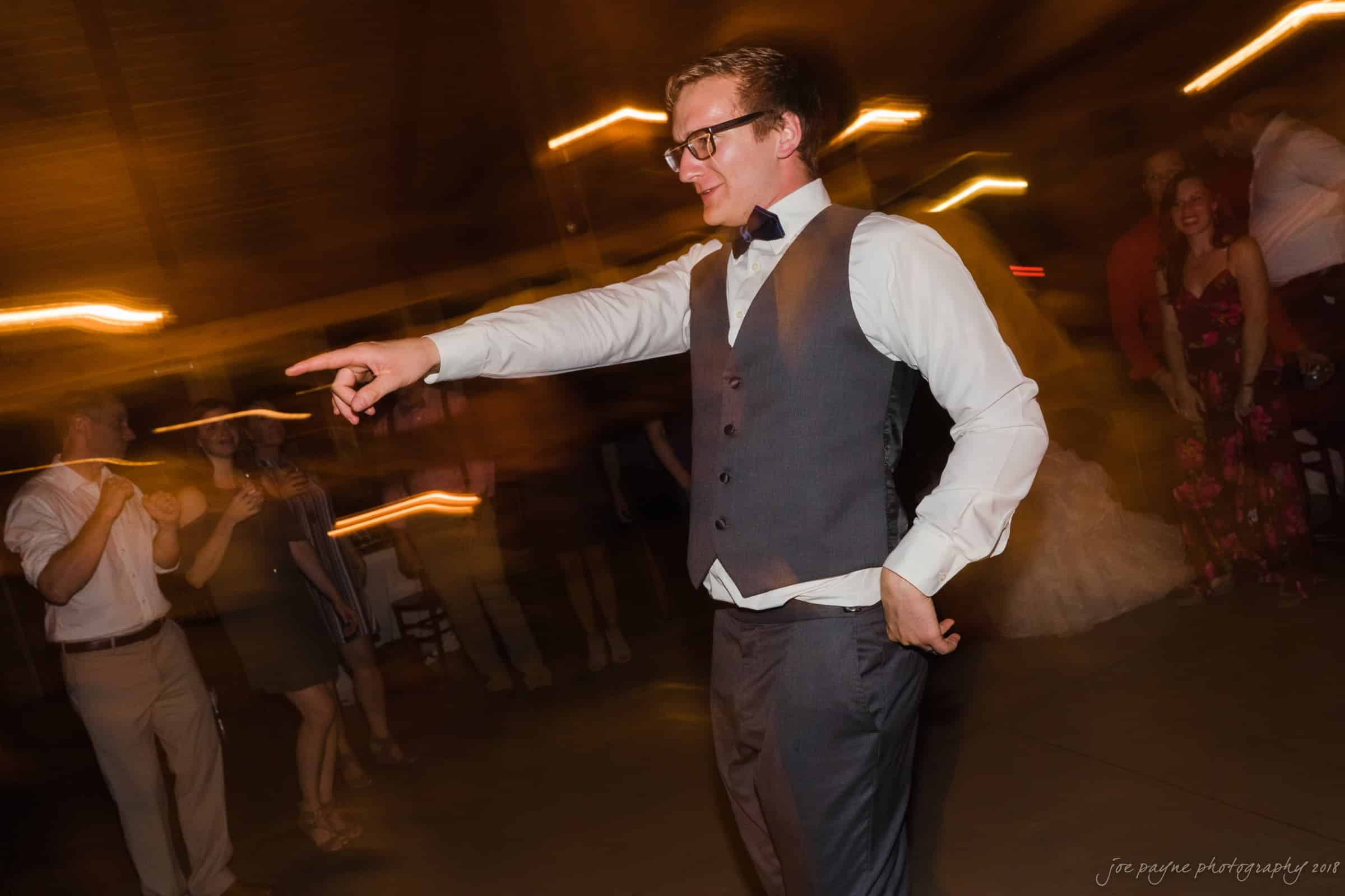 pavilion at carriage farm wedding - ashley & devon A person in a suit and bow tie energetically dances and points at a wedding reception. The background is filled with blurred lights and guests, creating a lively atmosphere.