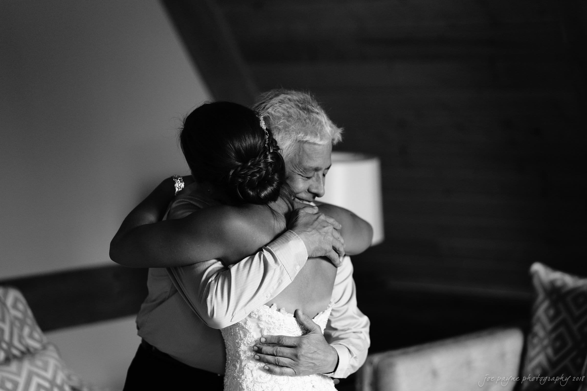 pavilion at carriage farm wedding - ashley & devon A black and white photo of two people embracing warmly. One person, wearing a wedding dress, faces away from the camera. The other person, an older individual with short hair, smiles as they share the hug. Wooden interior details are visible.